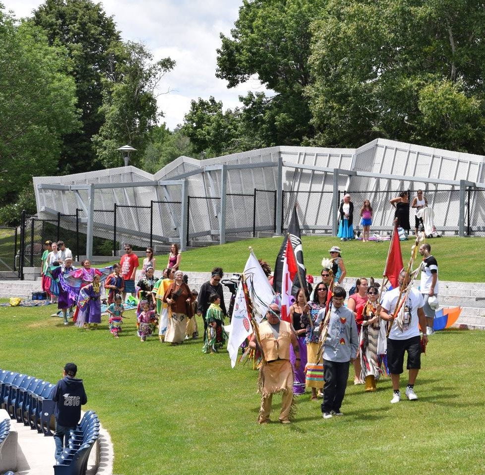 Dancers enter Bell Park in a line at the N'Swakamok National Indigenous People’s Day Pow Wow as people watch from off side. The grass and trees around them are green.