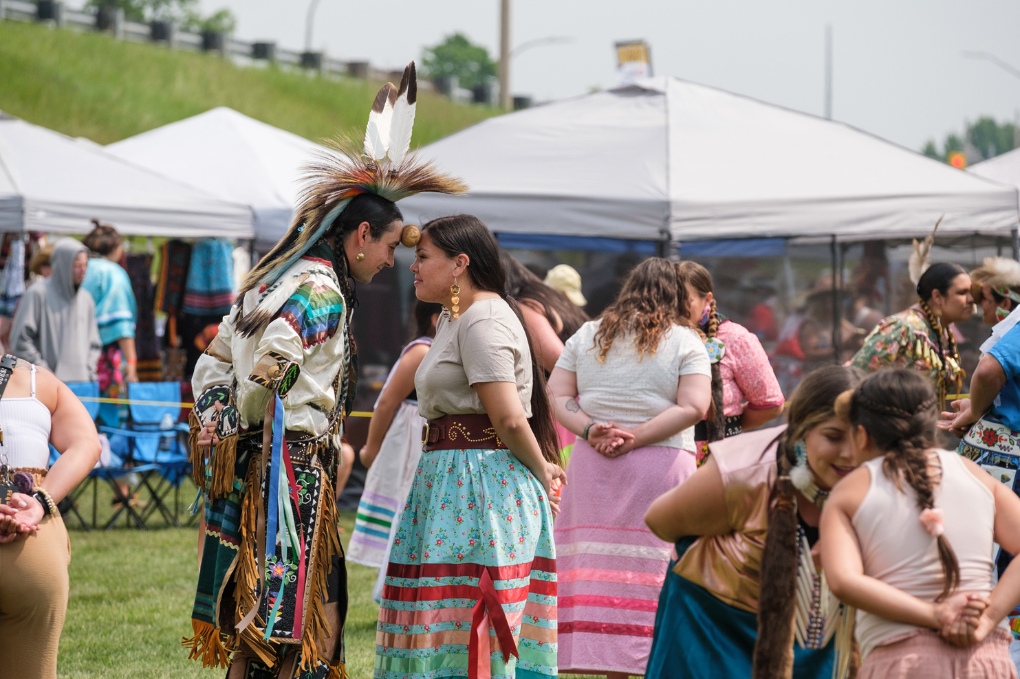 pairs of participants in beautiful traditional pow wow clothing delicately balance potatoes between their's and their partner's foreheads as they dance the potato dance. The partners have their hands clasped behind their backs and are smiling at each other and seem to be having a lot of fun.