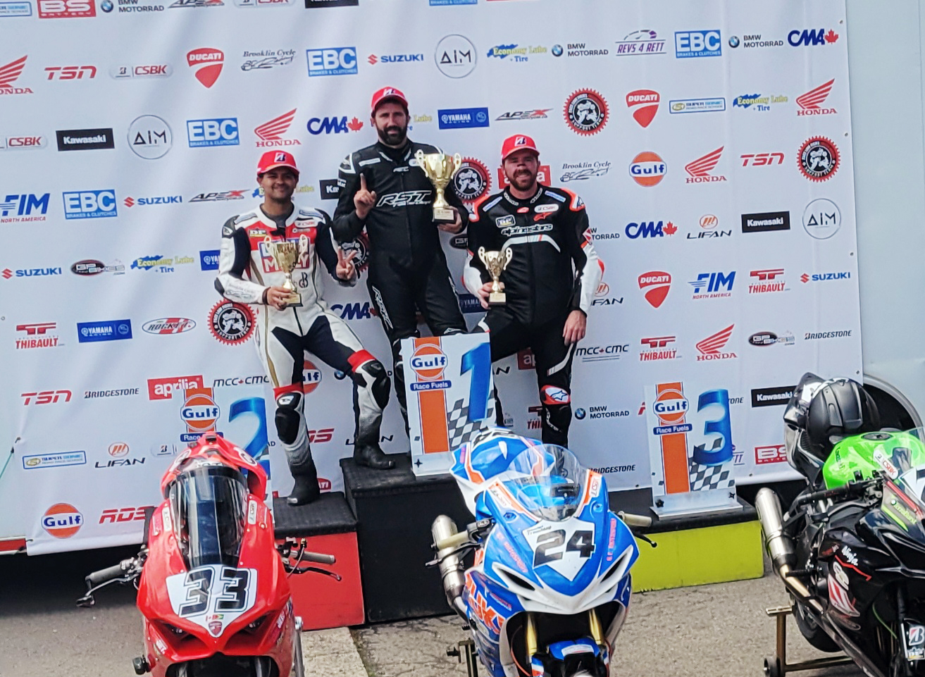 3 racers stand smiling on a low multi-level podium behind their parked motorbikes, posing with their first, second and third place trophies. 