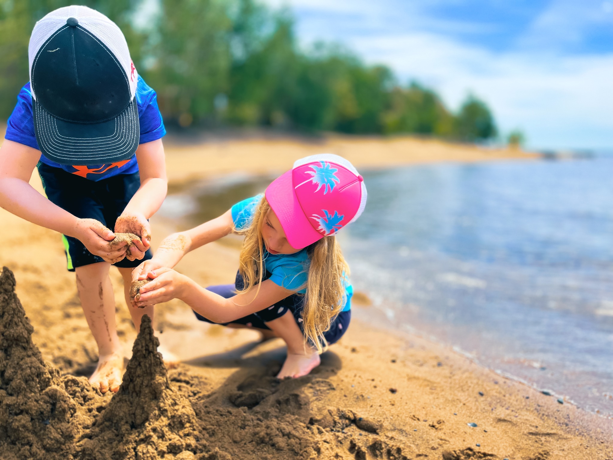 two young children building a sand castle in the soft sand of Pointe Des Chenes beach. Gentle sparkling waves of Lake Superior lap up just behind them. Green forest and blue sky are in the background. 