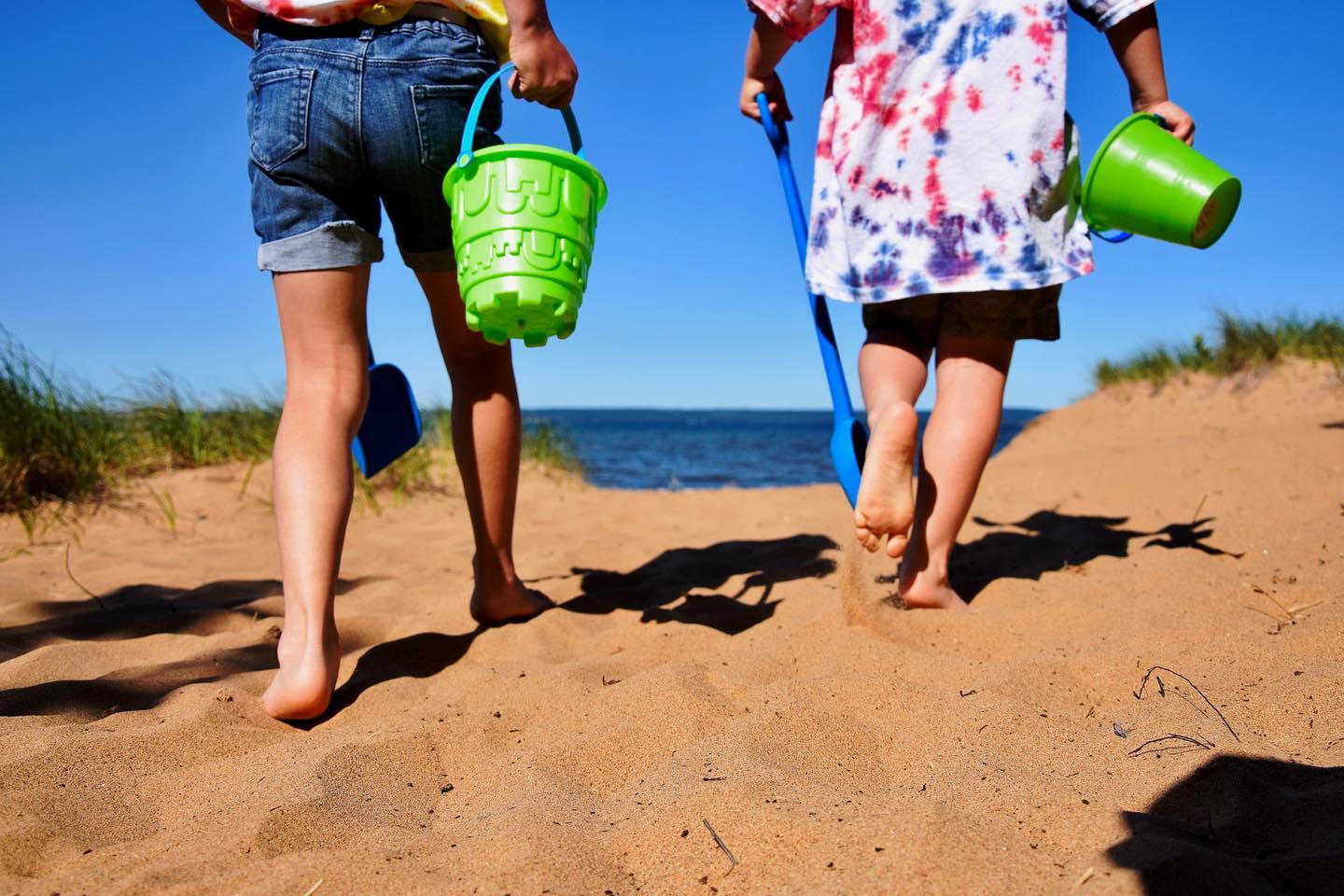 Two kids in shorts and carrying sand buckets walking through the soft sand toward the blue water of Lake Superior. The sky is a vivid blue and cloudless.