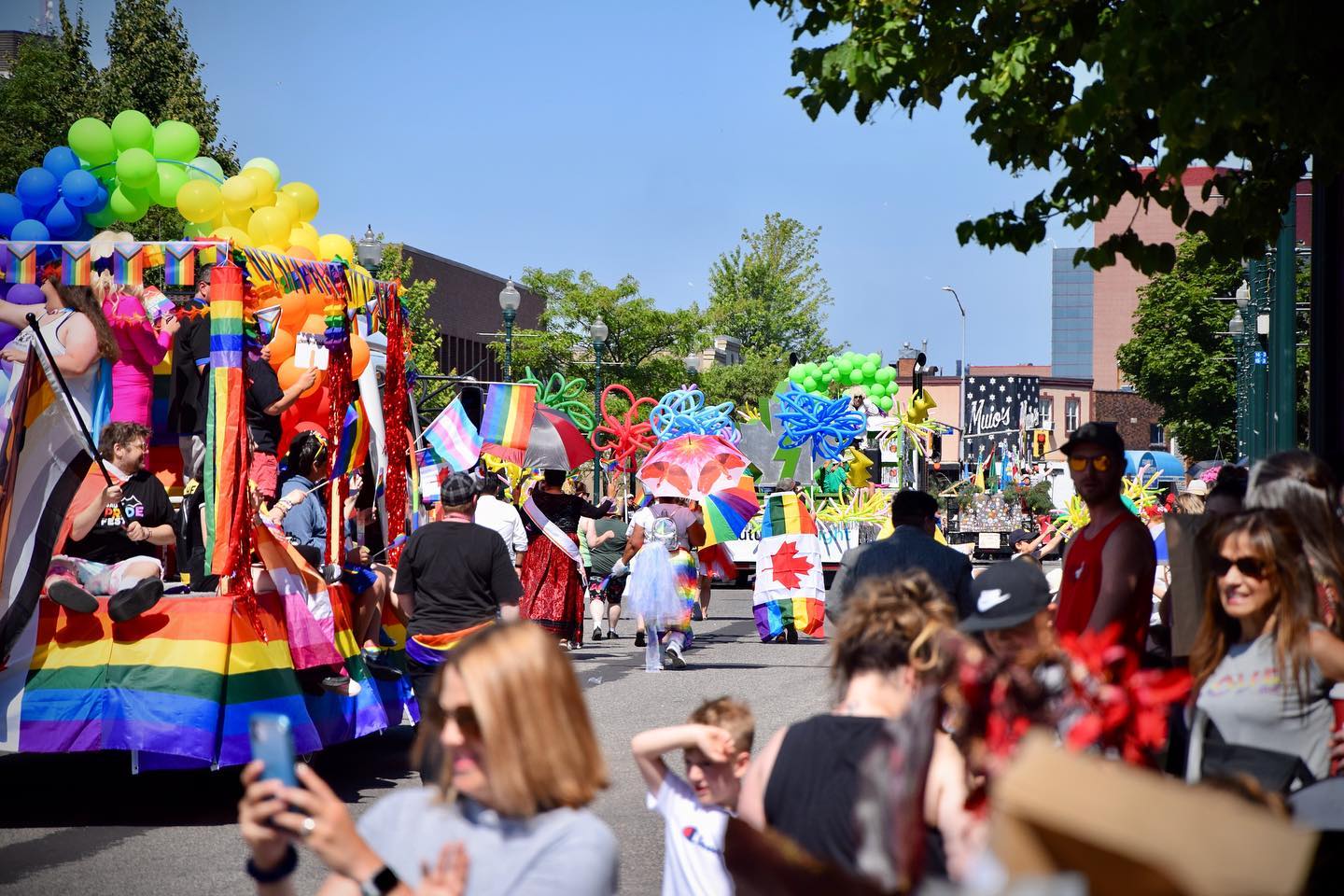 parade marchers and floats full of people, all decorated with rainbow balloons and banners, moving in a line down a Sault Ste. Marie street on a sunny summer day. A smiling crowd watches and takes photos. 