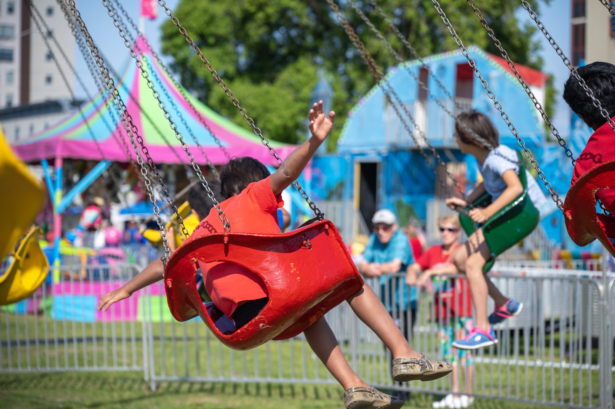 children ride an amusement park swing ride with their arms stretched out like wings as they swing high in broad circles while sitting in small bucket seats. A carousel, green trees, blue sky and carnival booths are in the background. 