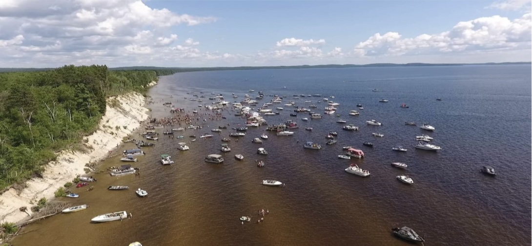 A large crowd of small boats anchored around a sandbank on Lake Wanapitei on a summer day. The water is calm and clear and there is thick green foliage on the top of the long sandy bank.