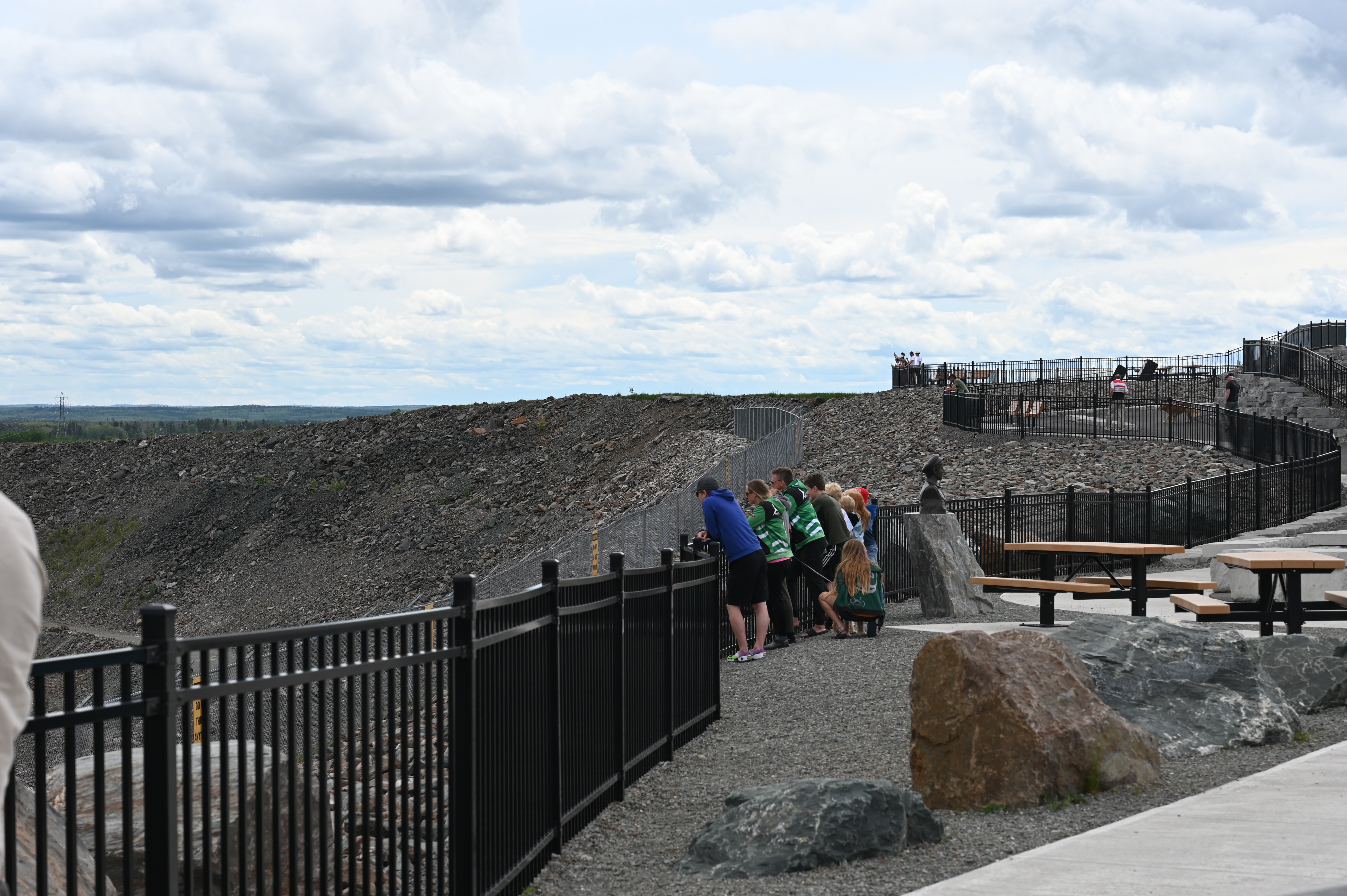 Zoomed-out side view of the Benny Hollinger Bust at the Hollinger Open Pit Lookout in Timmins with people standing behind it 