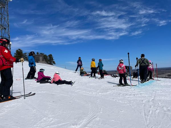 Peoples dressed in warm clothing are standing and sitting at the top of the ski hill and are holding their skis