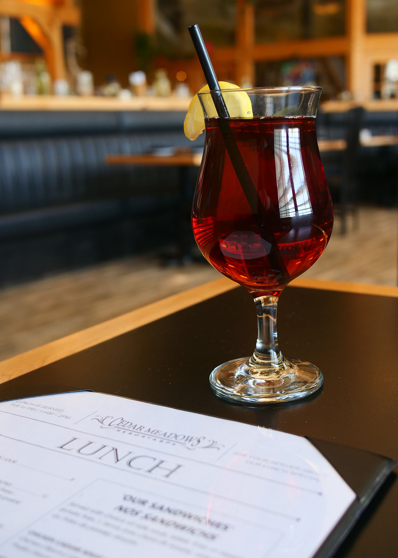 A red cocktail with a straw and lemon wedge, on a black table next to a Cedar Meadows menu, with a dark wood bar in the background. 