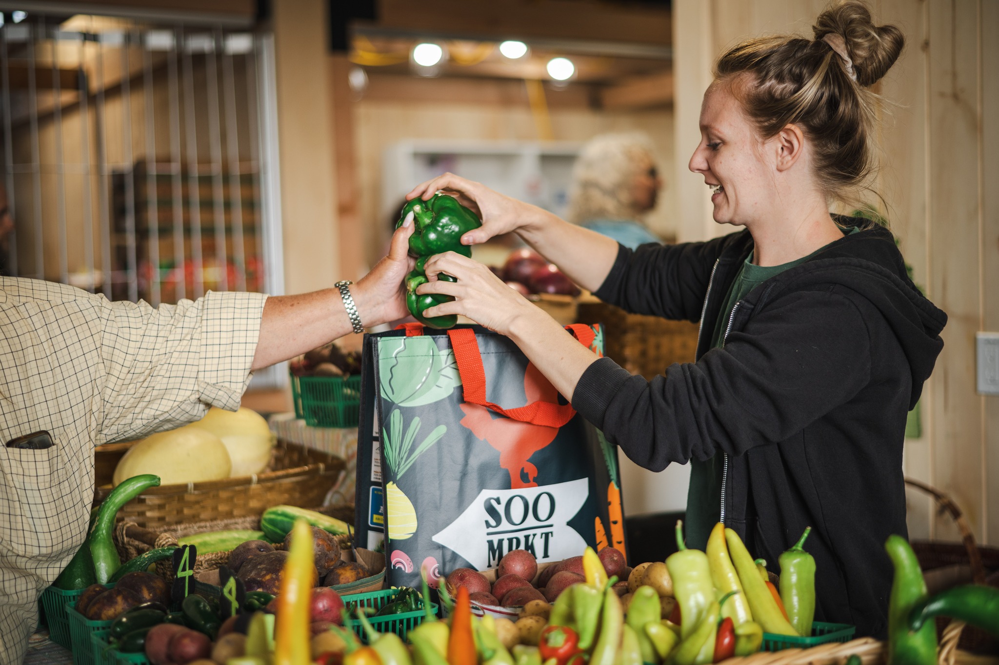 A smiling vendor at a table full of colourful fresh produce helps a customer to put some vegetables into their bag, which is labelled "Soo Market".