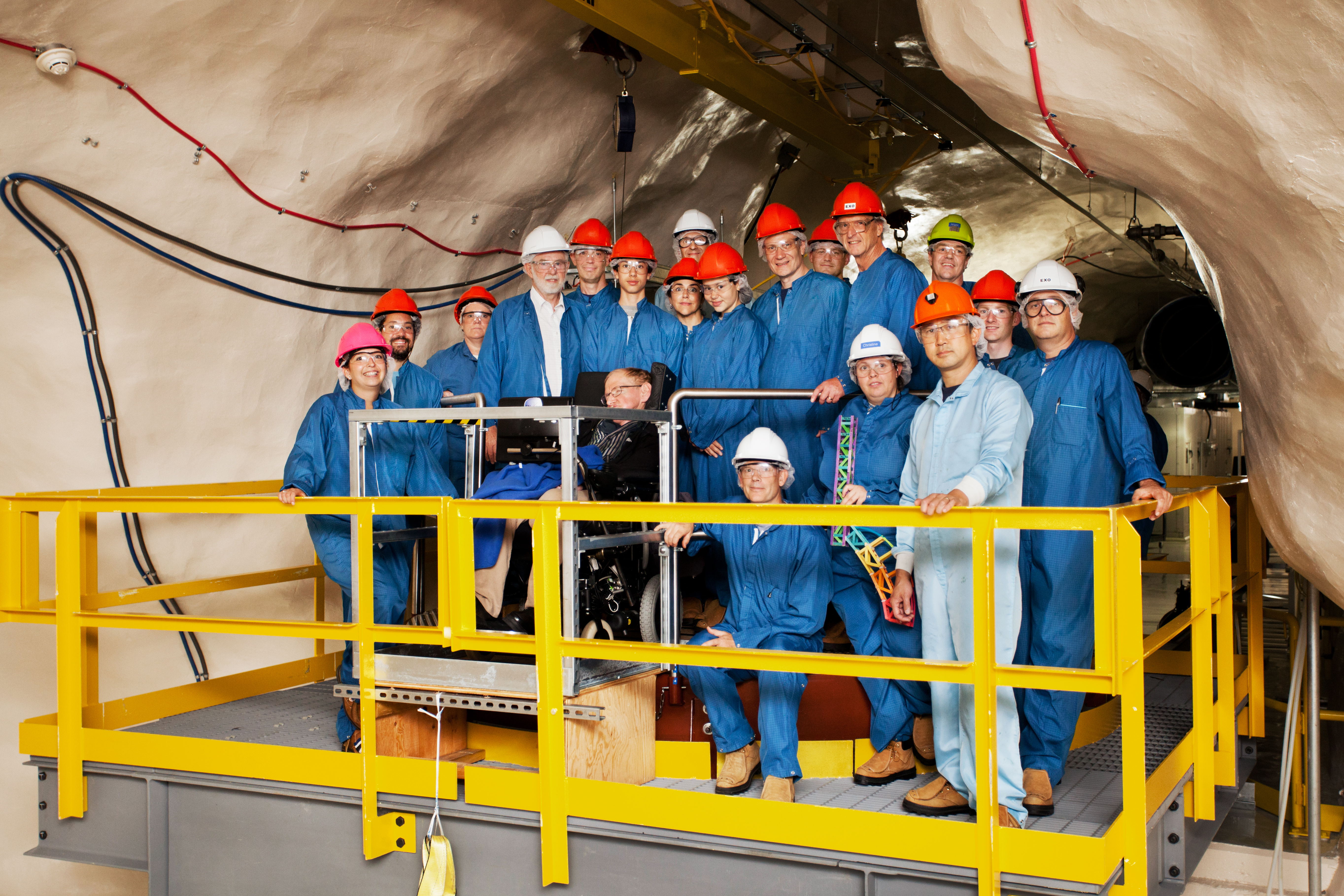 Scientist Stephen Hawking in a wheelchair surrounded by workers in blue coveralls and orange construction hats.