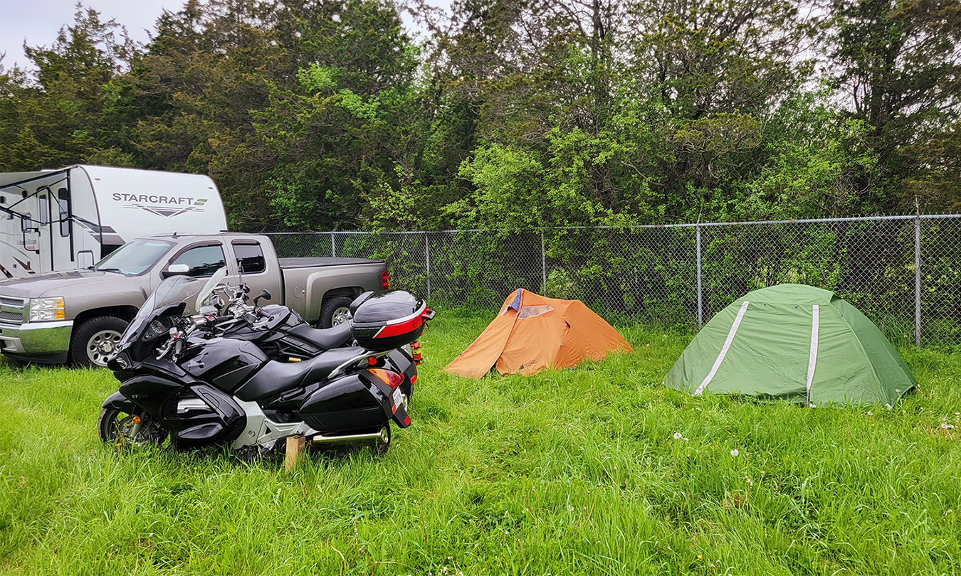 two small tents set up next to a camper and a motorbike in a field of knee-length, very green grass. Behind them is a chainlink fence and green forest.