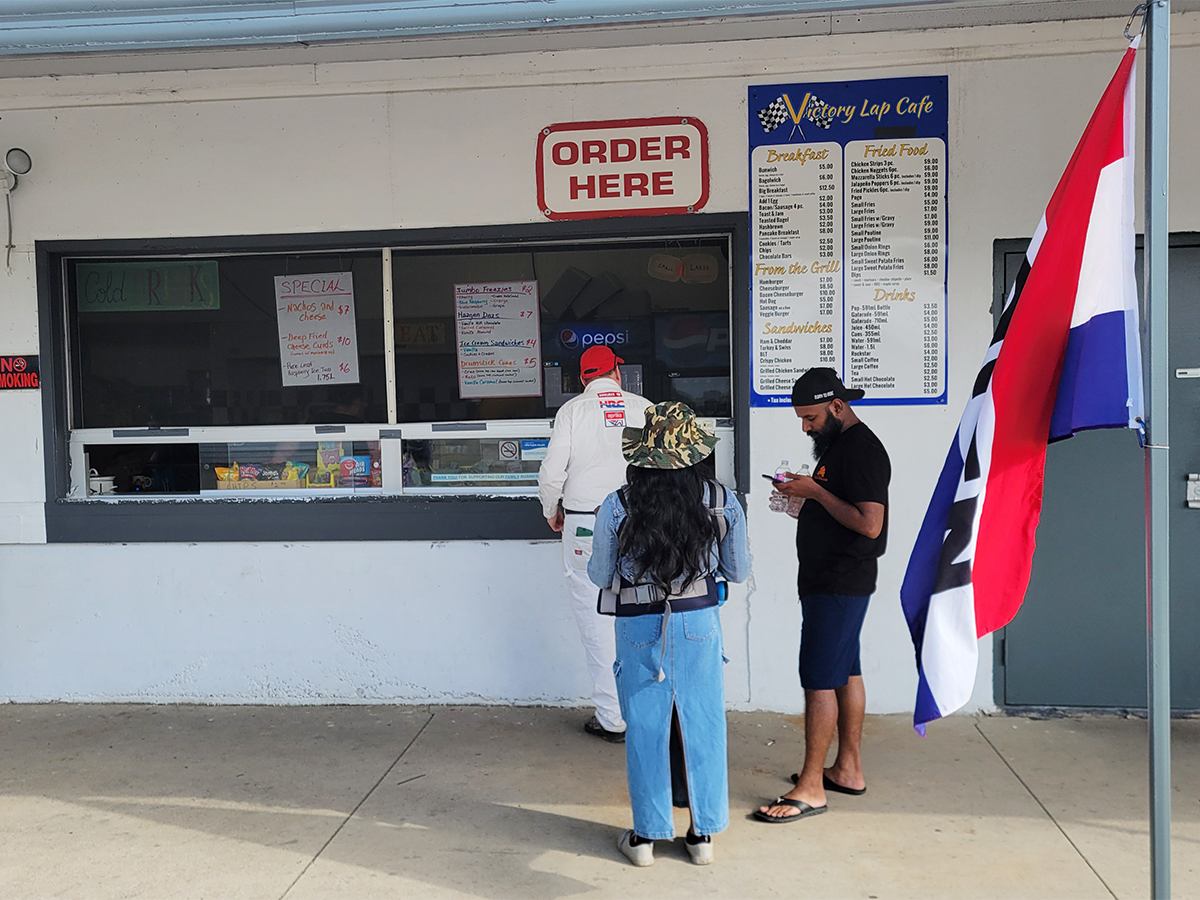 3 people standing in line at a concession booth window at a Superbike event. One is ordering at the window under a sign that says "order here". A menu is posted on the wall of the concession that reads "Victory Lap Cafe", and lists breakfast foods, sandwiches, grilled and fried foods. 