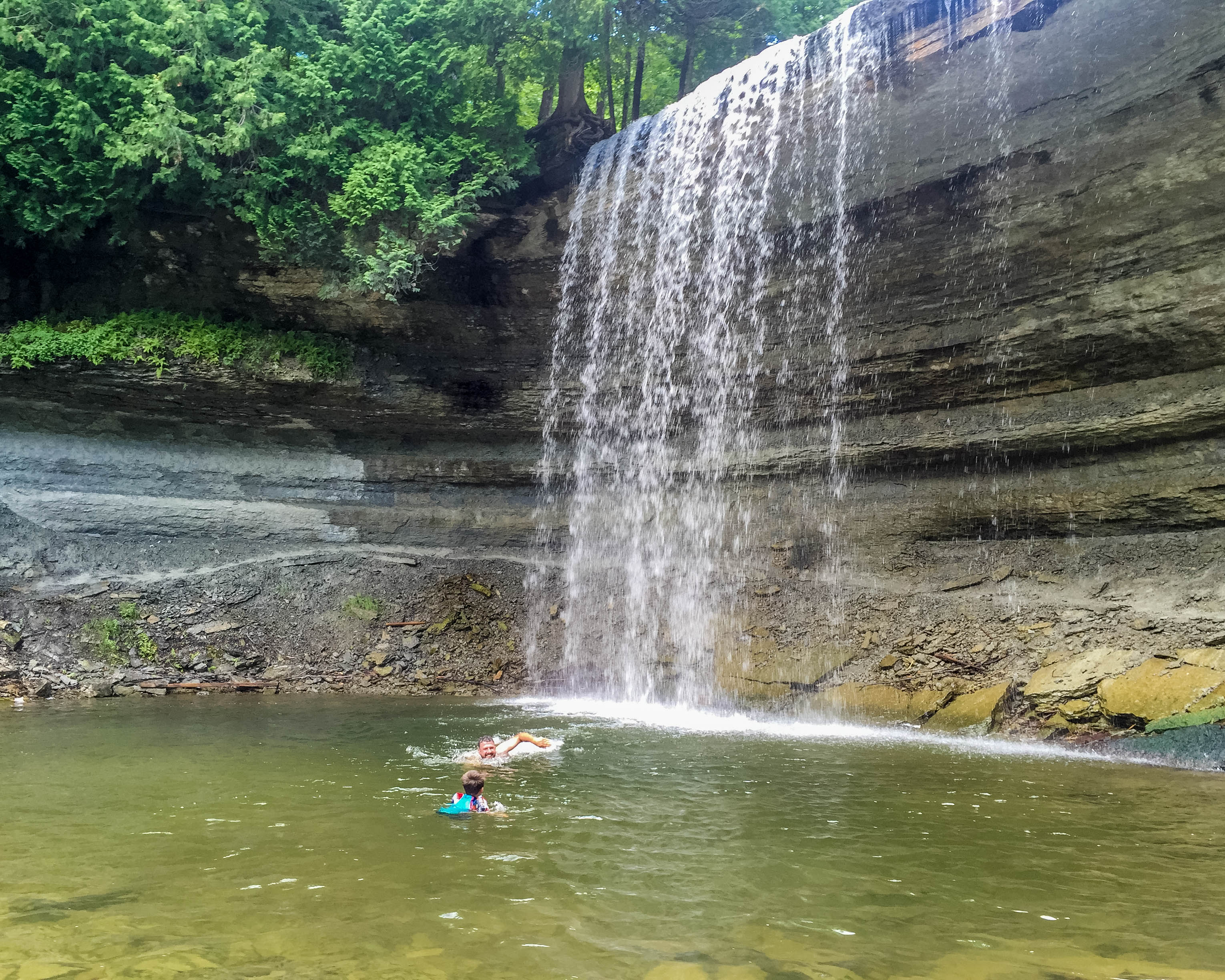 A father and son swim in the calm clear pool at the bottom of Bridal Veil Falls in the summer.