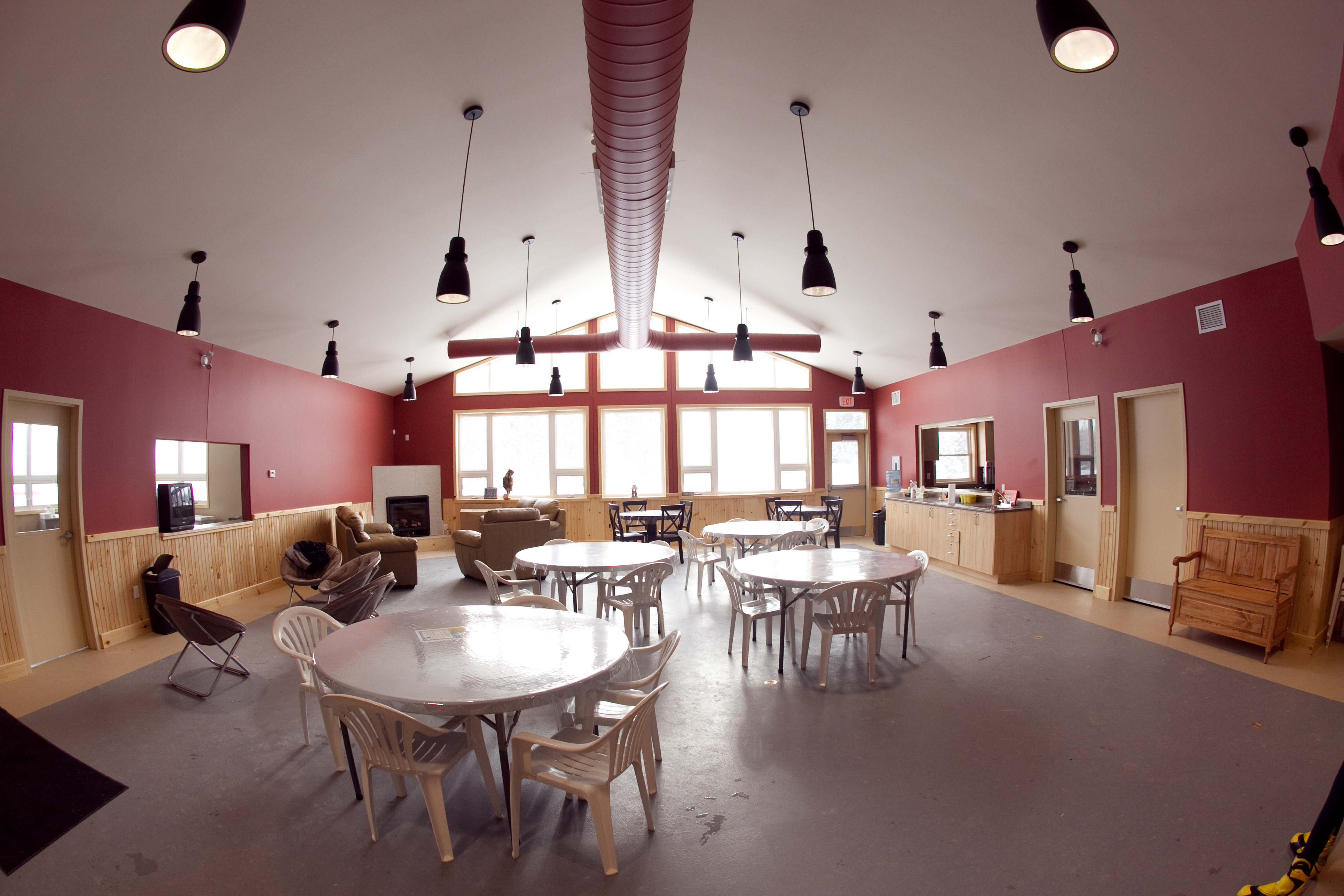 a wide view of the inside of the Porcupine Ski Runners lodge which features red walls and approximately 4 tables and chairs in the middle of the room, a fireplace on the left corner and some benches on either side of the walls