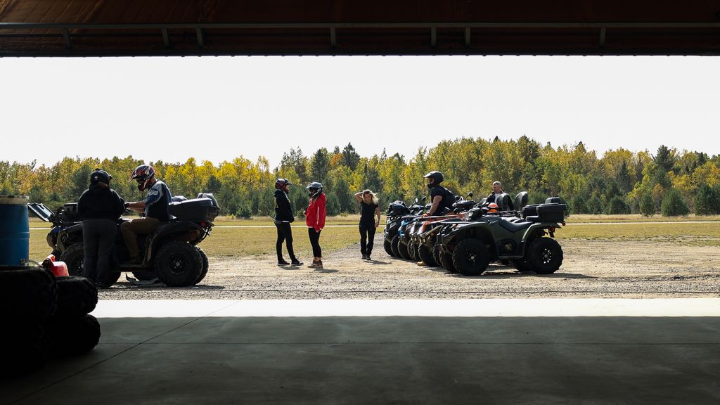 a group of people stand outside a garage, talking and readying a row of ATVs for a ride. The day is sunny and there is green forest in the background.