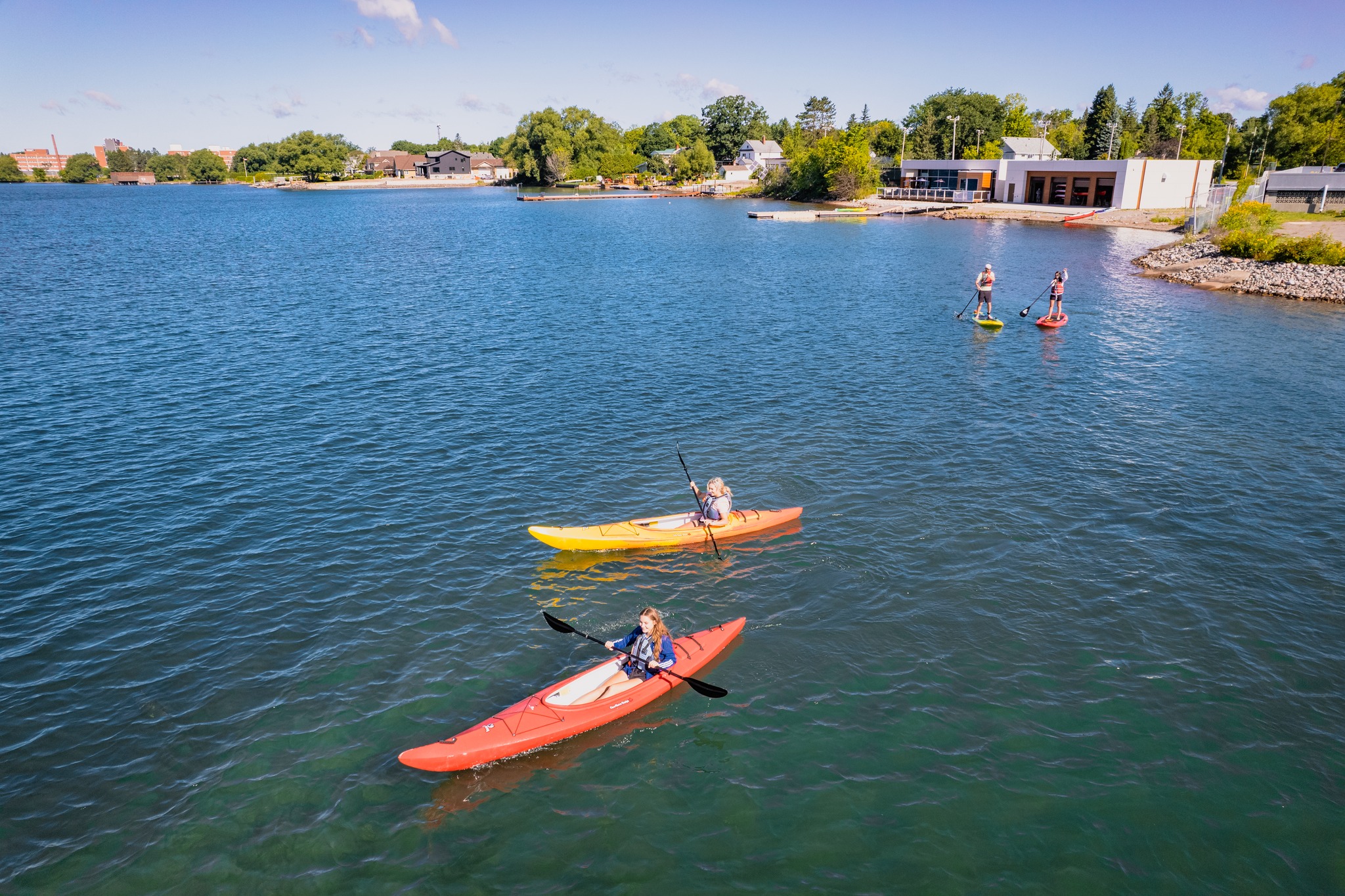 2 kayakers and 2 paddleboarders paddle out from shore on the turquoise water of Lake Superior in the summer. The pristine shore is full of green trees and short buildings, and the sky is clear blue.