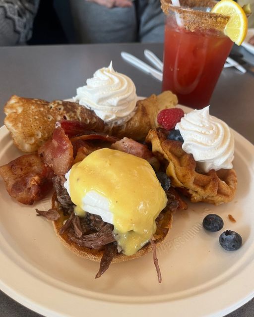 a plate containing eggs benedict, waffles, bacon and toast with a drink beside it