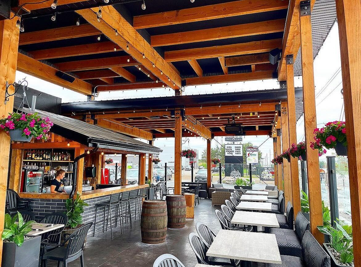 An outdoor patio at Gino's in Sault Ste. Marie, with rows of comfortable tables and chairs, thick wooden beams making up a roof for shade and a covered bar. There are wooden barrels and lots of potted plants and hanging flowers for decor.