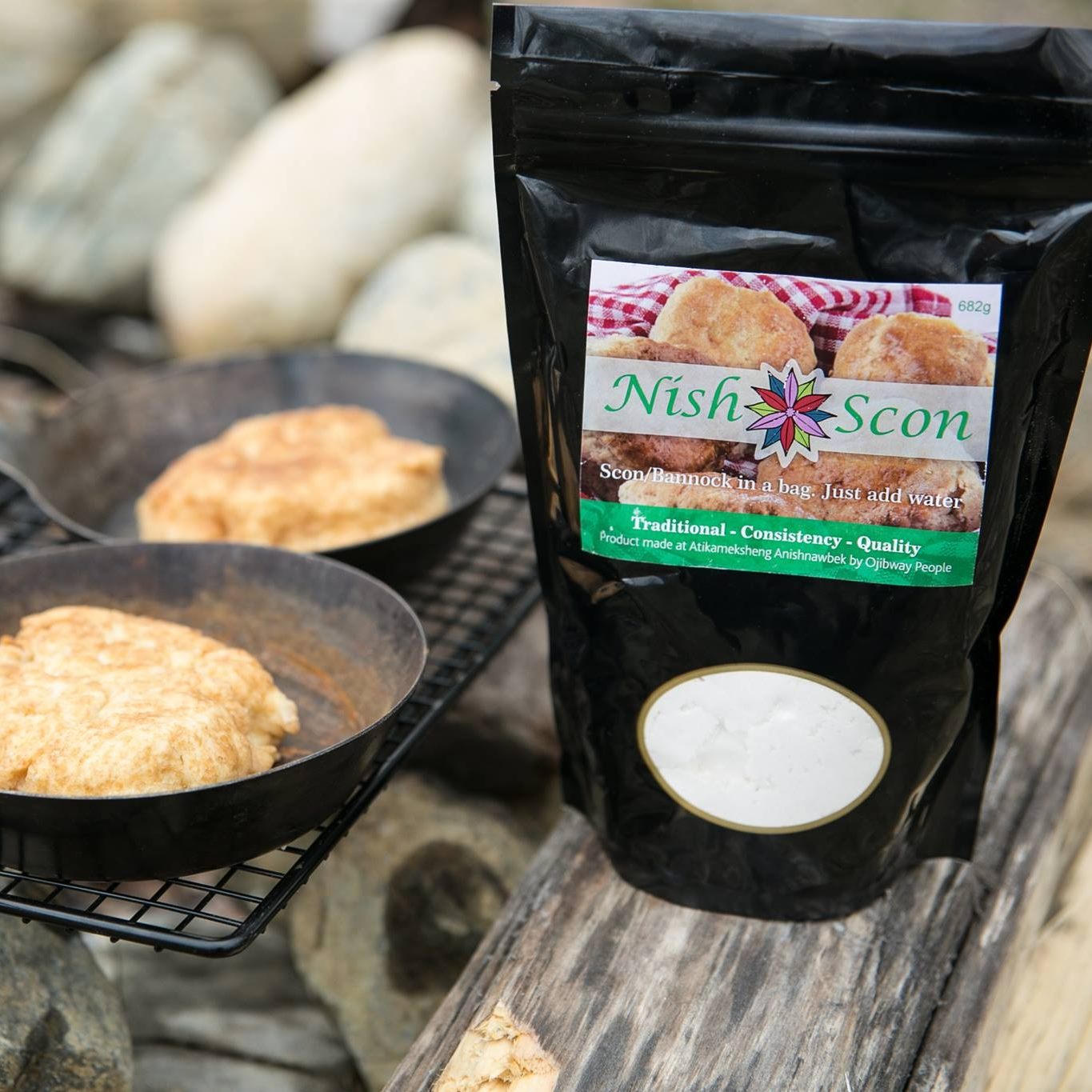 Two scons cooking over a fire in a cast iron pan alongside a black package of NISH brand scon mix. 