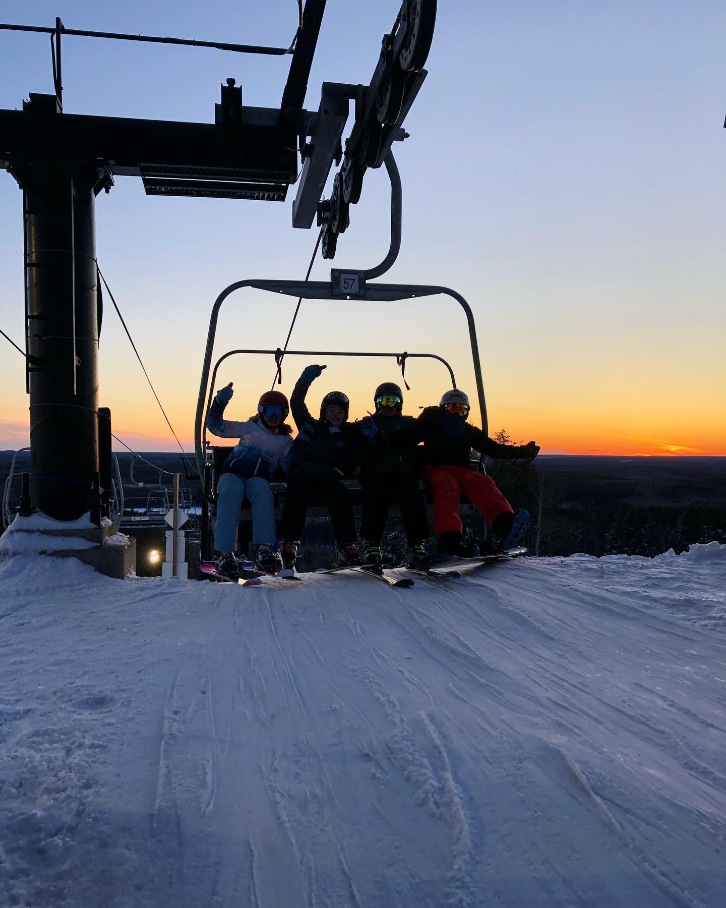 4 skiiers are dressed in their winter clothes and are wearing skis while riding a chair lift. Behind them is a sunset