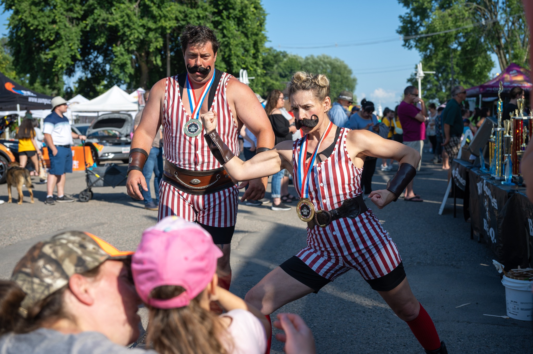 two actors wearing the striped short-and-tank body suits and black handlebar moustaches of 1920s carnival strongmen flex and entertain a group of onlookers at the Rotaryfest in Sault Ste. Marie.