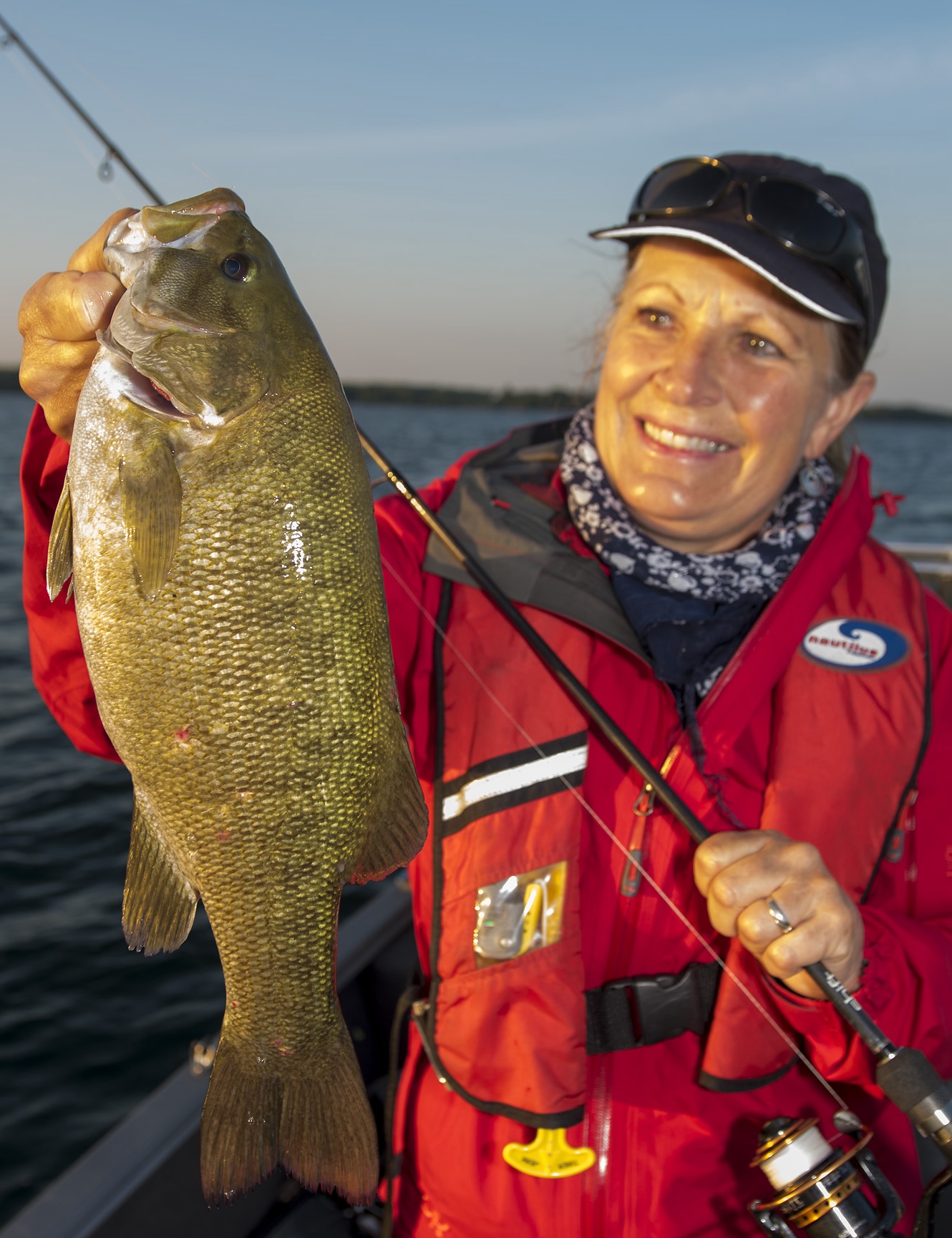 A smiling woman holding up a fishing rod and a large smallmouth bass. She is standing in a boat at sunset. 