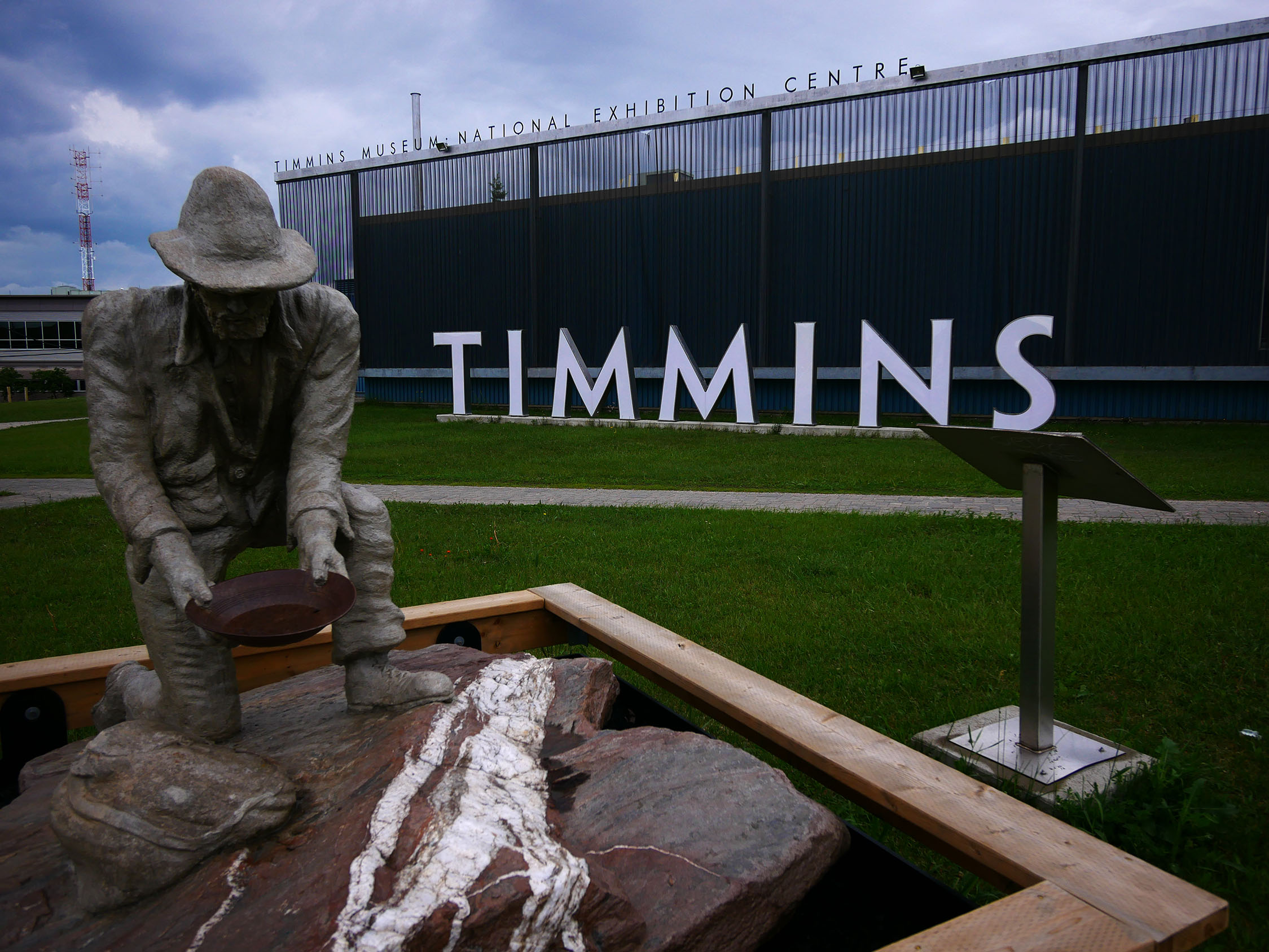 a detailed statue of a man crouching on one knee next to some rocks, panning for gold. Behind the statue is a green park fied and a sign of tall free-standing metal letters that spell "TIMMINS", under smaller lettering that says "Timmins Museum National Exhibition Centre".