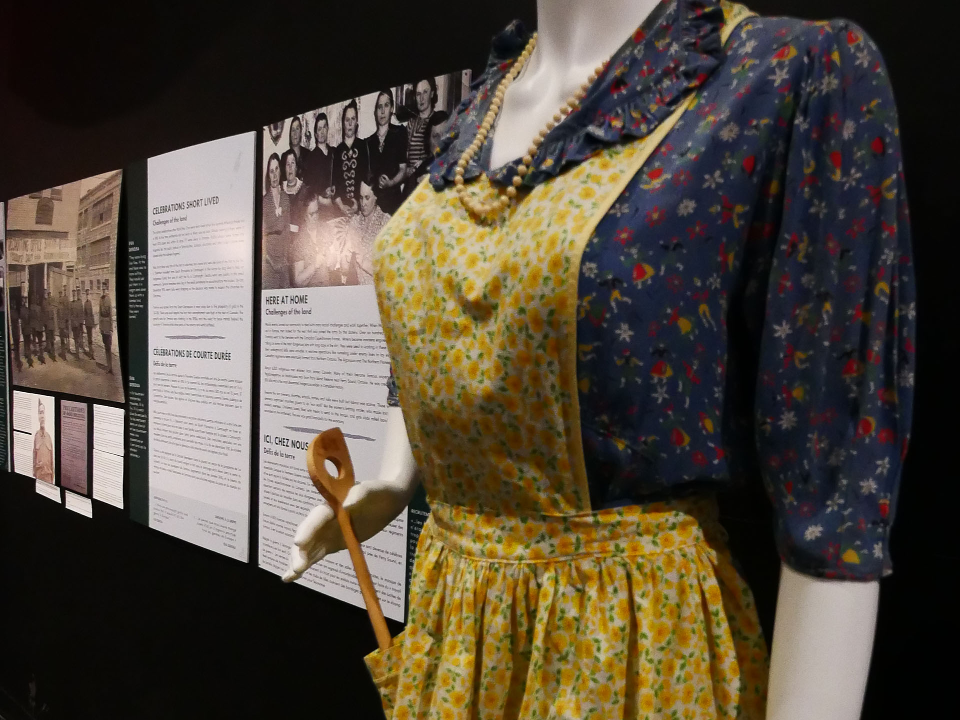 an antique blue flowered dress and beige flowered apron on a mannequin next to several illuminated interpretive panels about the history of the Timmins area.