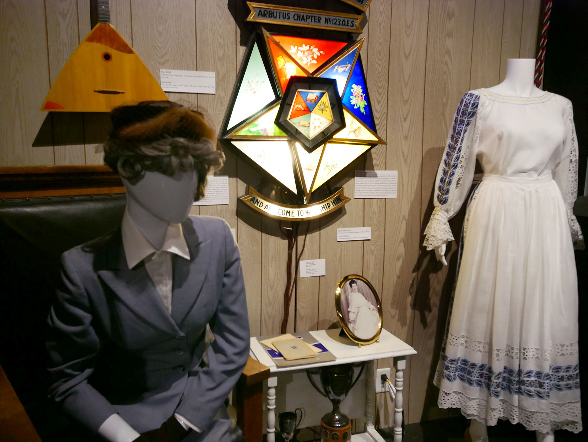 an antique grey suit and a white dress on mannequins, a stained glass lamp, an end table and a clock set up as part of a historical display at the Timmins Museum.