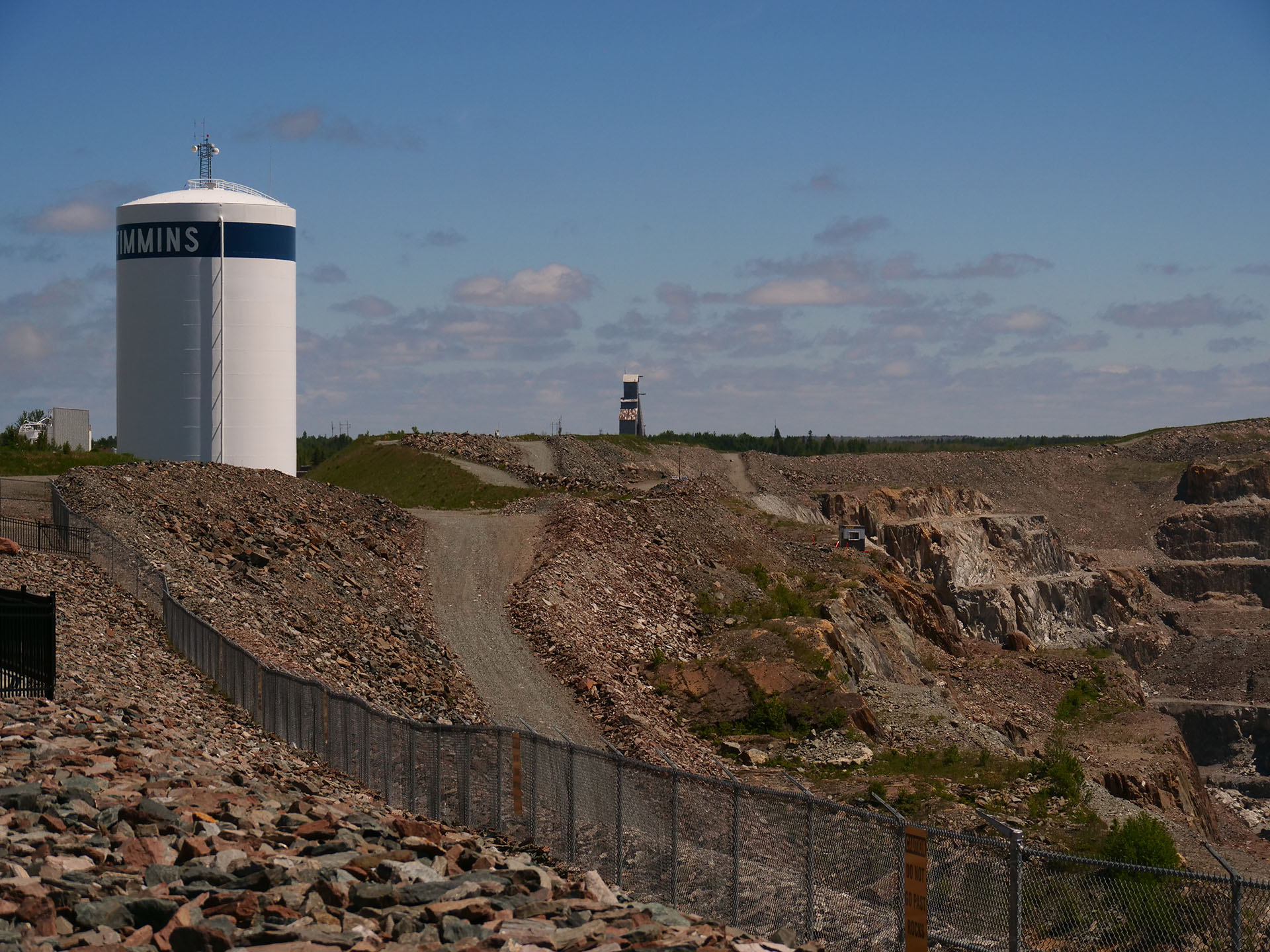 The rocky, sloping landscape of the Hollinger Mine pit area, edged by a long chain link fence. The headframe is on the horizon.