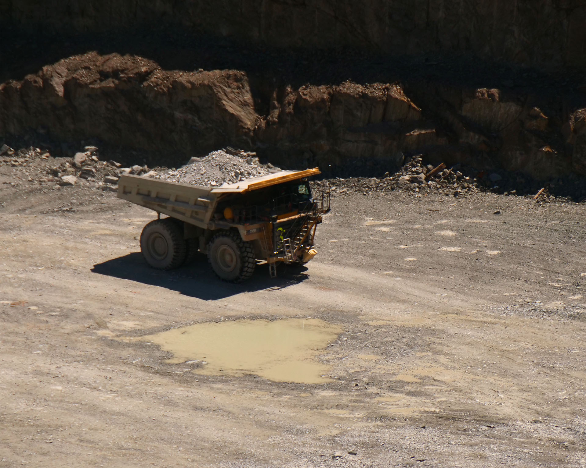  a huge mining truck with the back filled with rocks drives along the bottom of the mine pit.