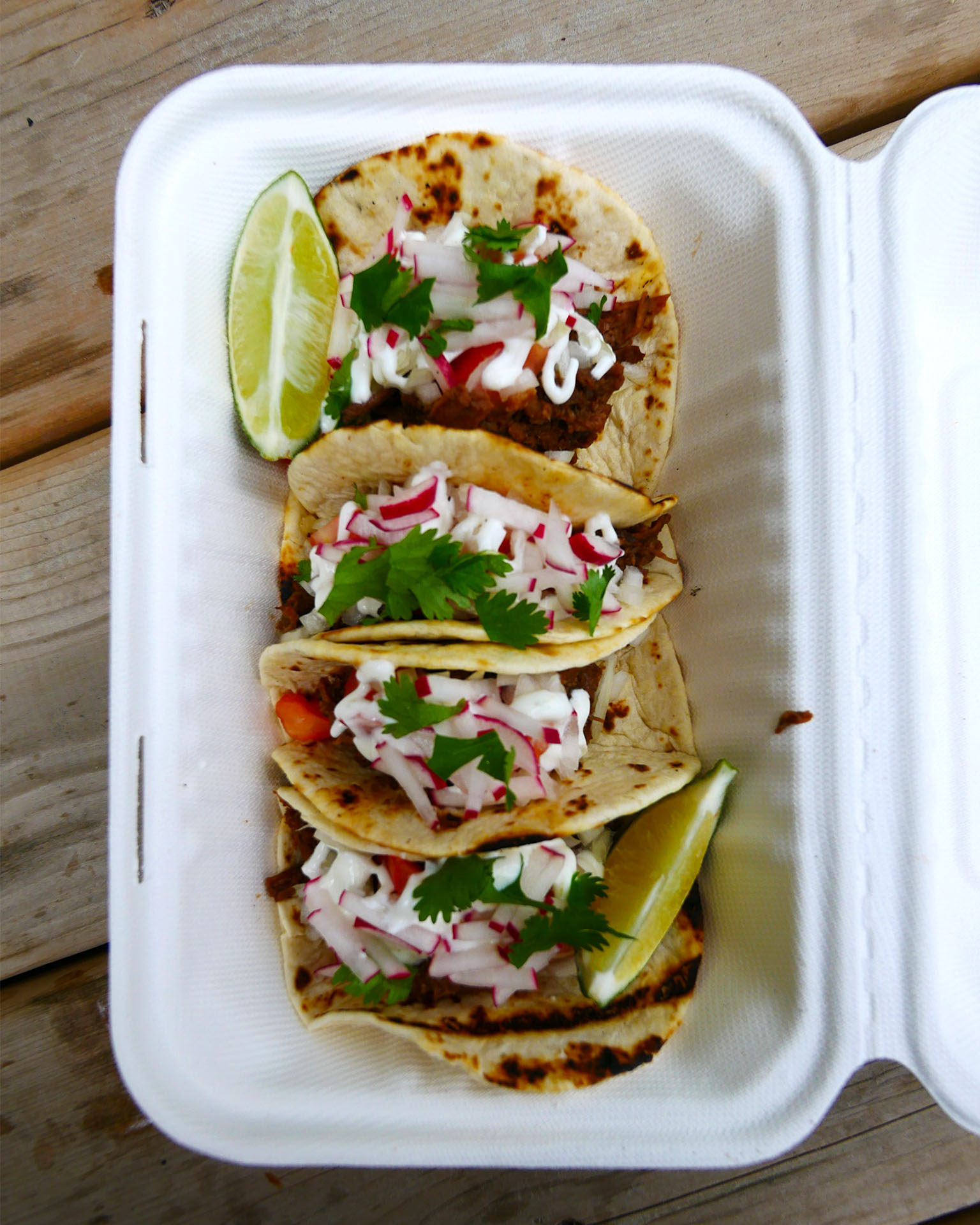 A take-out container of 4 fresh beef tacos on yellow corn tortillas topped with shredded radish, cilantro and lime.