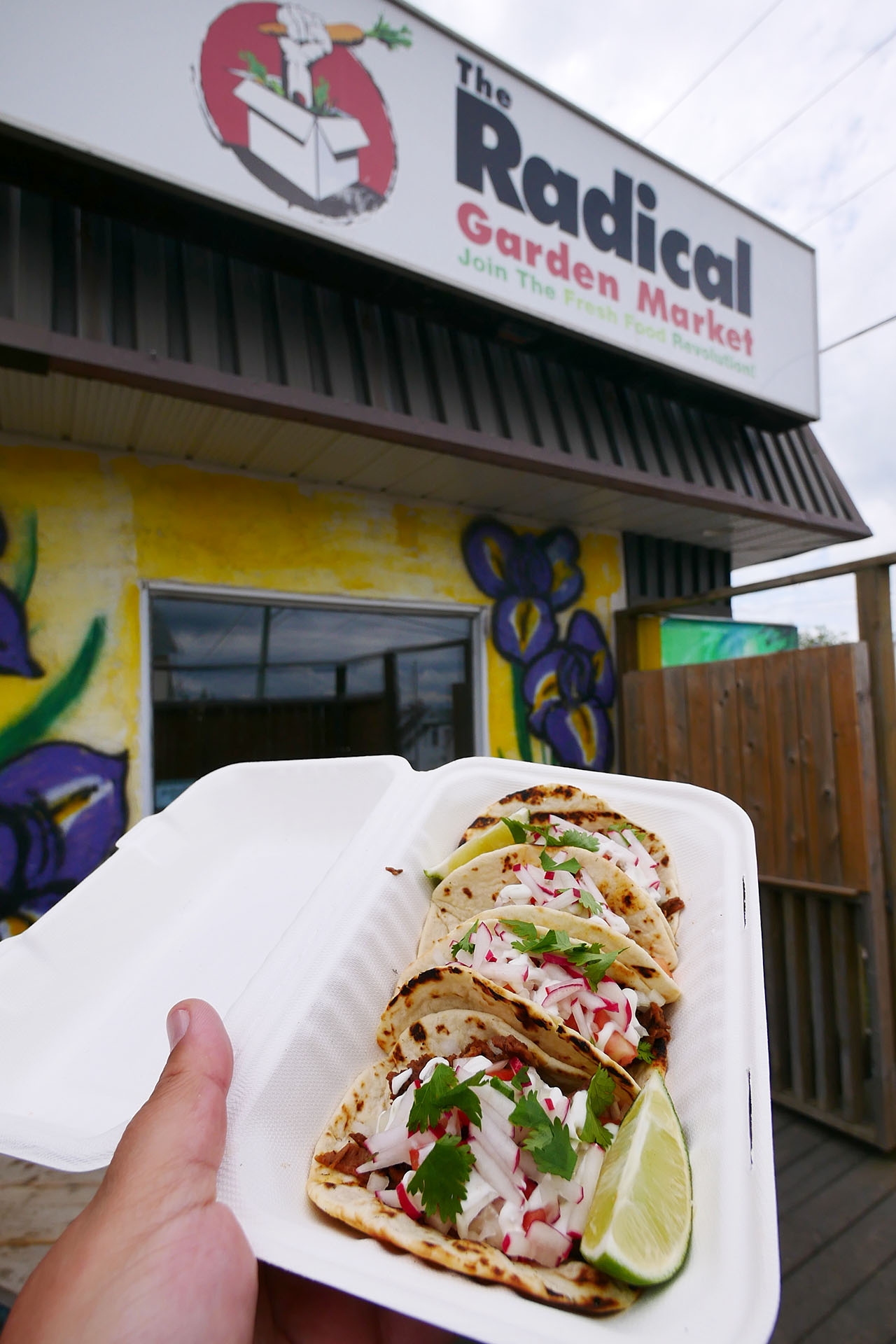 a hand holding up a container of tacos outdoors in front of the Radical Garden Market sign. 