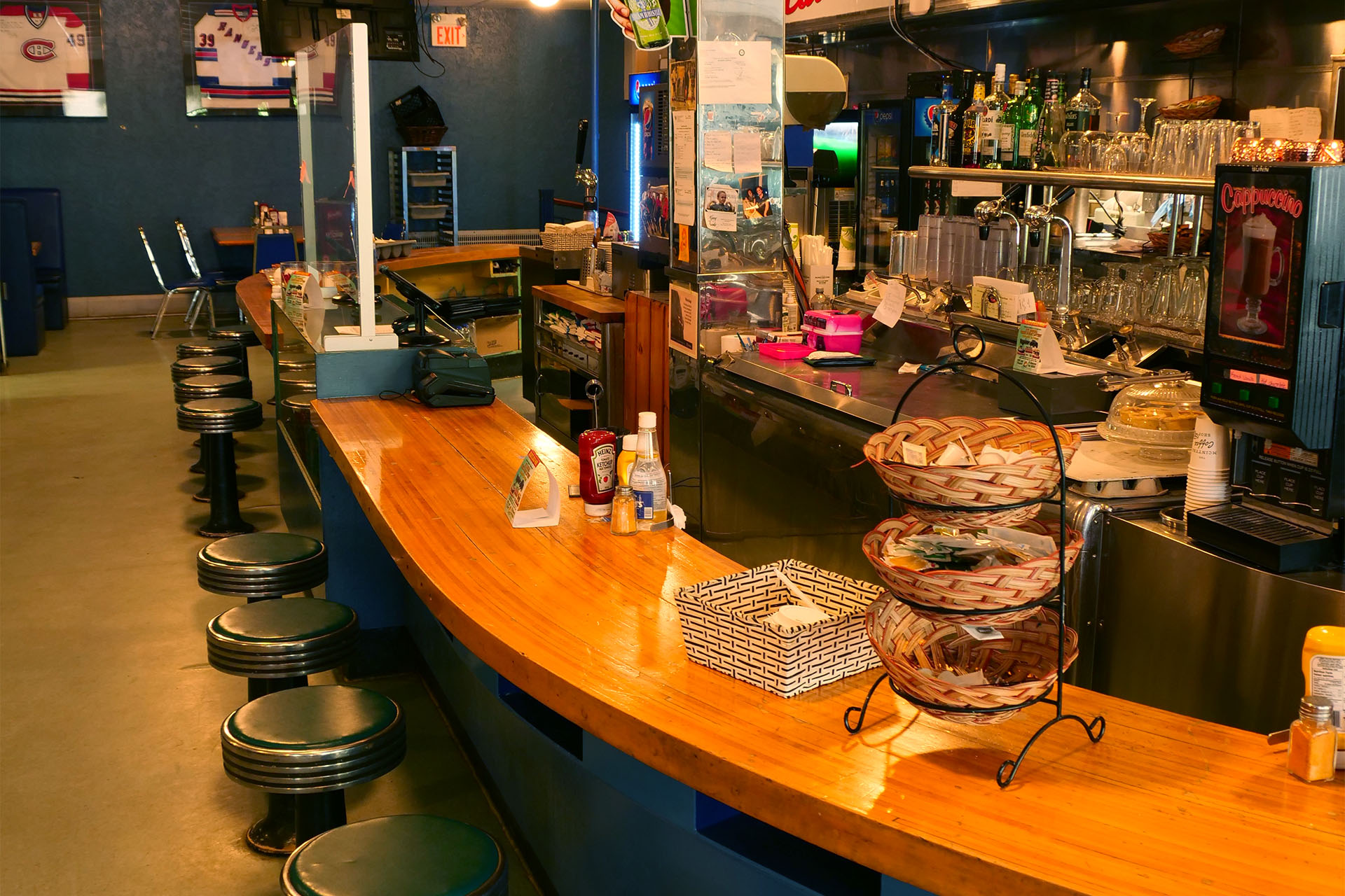 The inside of the McIntyre Coffee Shop; a long wooden front counter with stools for customers, pies in a display rack on the counter and coffee machines and dishes behind it. The coffee shop is designed to look and feel cozy and old-fashioned. 