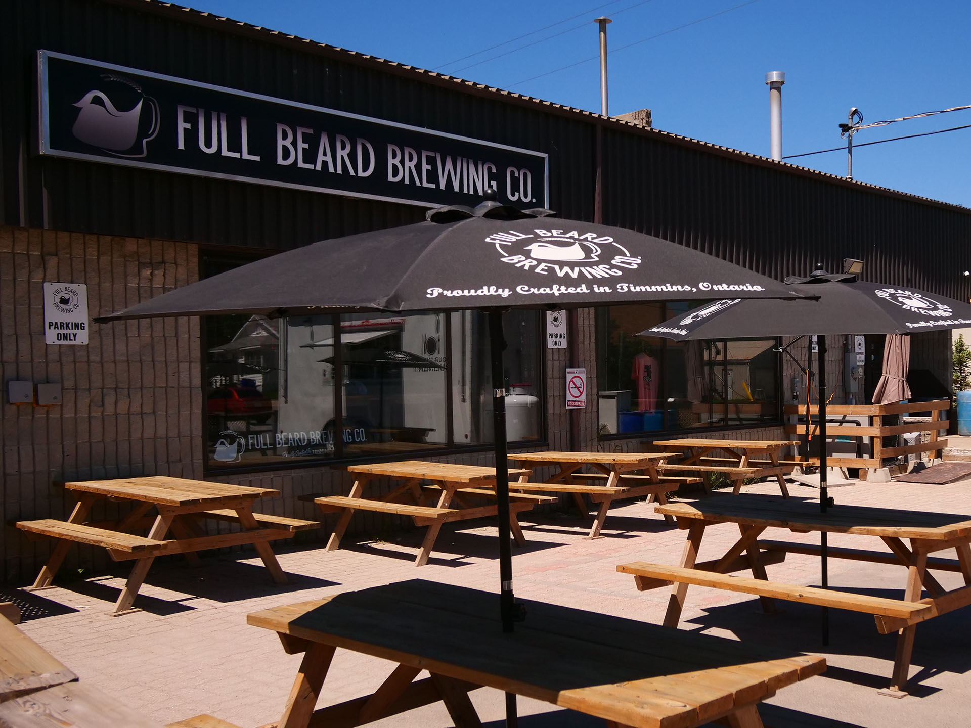 An outdoor patio at Full Beard Brewing in Timmins; two rows of wooden picnic tables with black umbrellas labelled with the company logo.