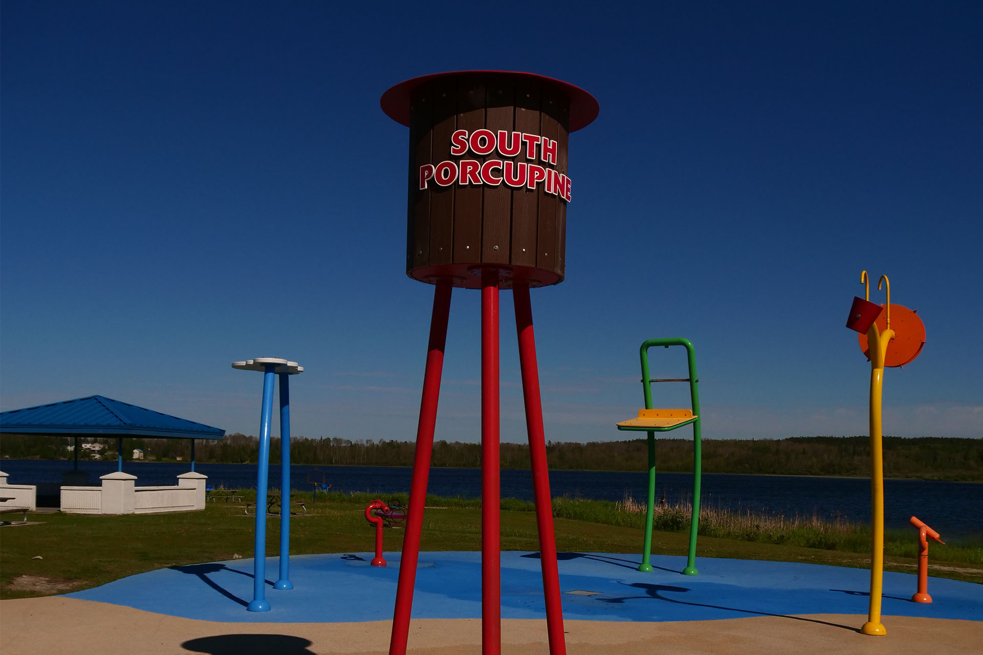 a spray park next to a gazebo in a large green field under a  deep blue, cloudless summer sky. One of the spray structures in the park is shaped like a wooden watertower, and labelled "South Porcupine" in large red letters.