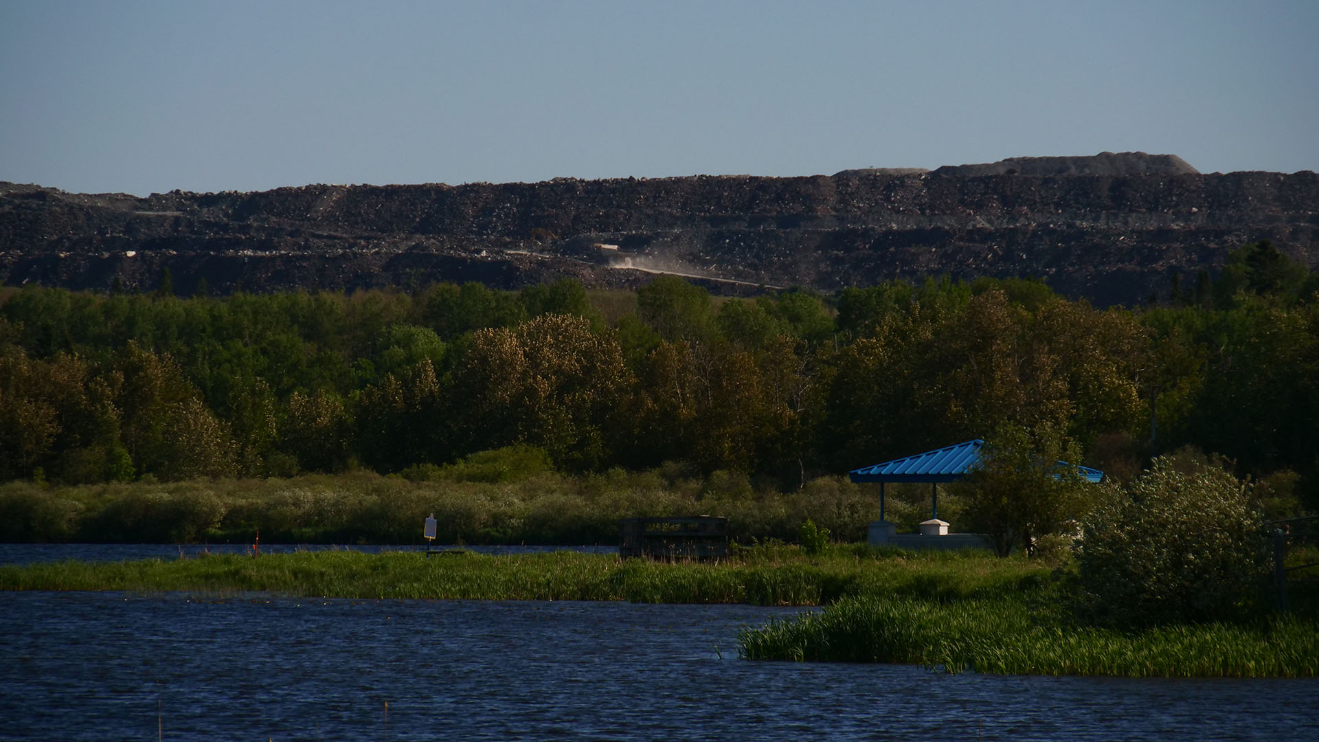 White Waterfront on a summer day; an expanse of deep blue lake surrounded by green forest, with a blue roofed gazebo on one edge. In the distance a cloud of dust is raised by a large mining truck heading up a road on the horizon.