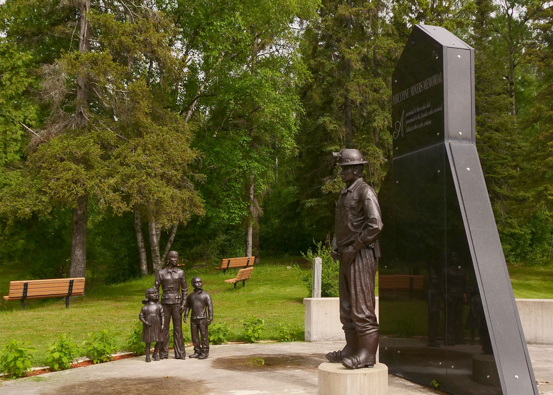 A display in Lions Park, with a black granite stone cut to the shape of the McIntyre Headrame next to a bronze statue of a man in vest and hat. Further in the background is another bronze statue of a man standing with two children. Green trees and grass and two wooden benches are in the background. 