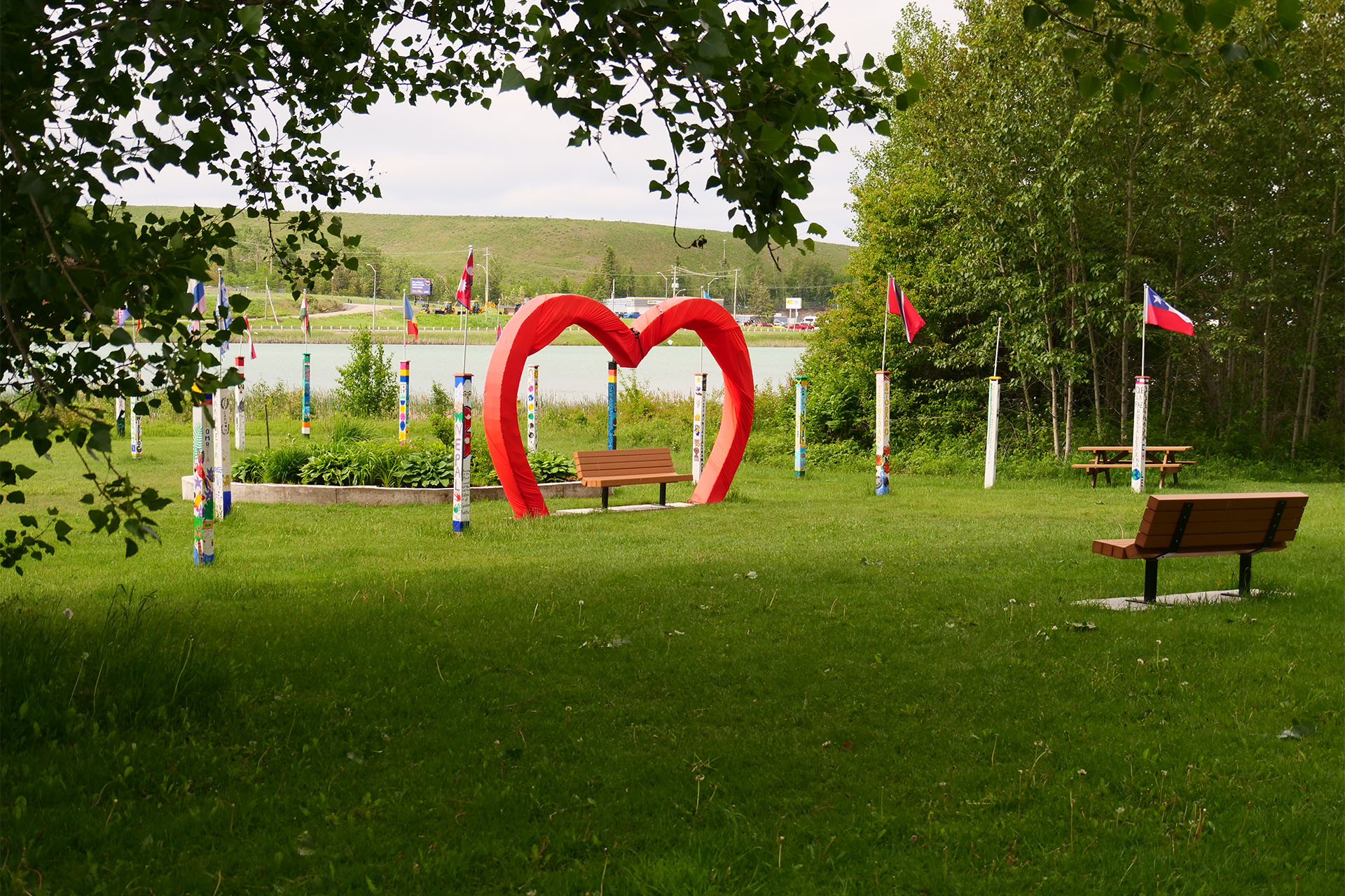 Lions Park in Timmins; a very green grassy space surrounded by tall shade trees and dotted with wooden benches with a lake in the distance. One wooden bench has a red metal structure arching out of the ground and over the bench in a heart shape. 