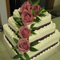 a close up of a four-tier square white wedding cake with pink roses that go down one of the corners and three roses on top. There is a piped design along the top of the edges and brown or dark red on the bottom of all the tiers