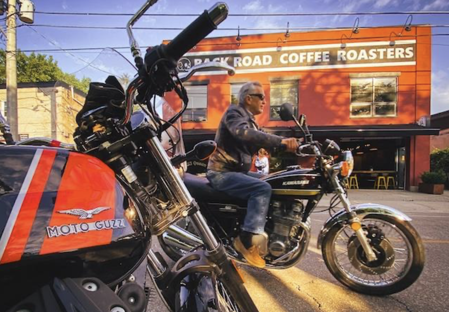 two motorcyclists ride down the street in front of a red shop building bearing the sign "Back Road Coffee Roasters" on a summer morning.