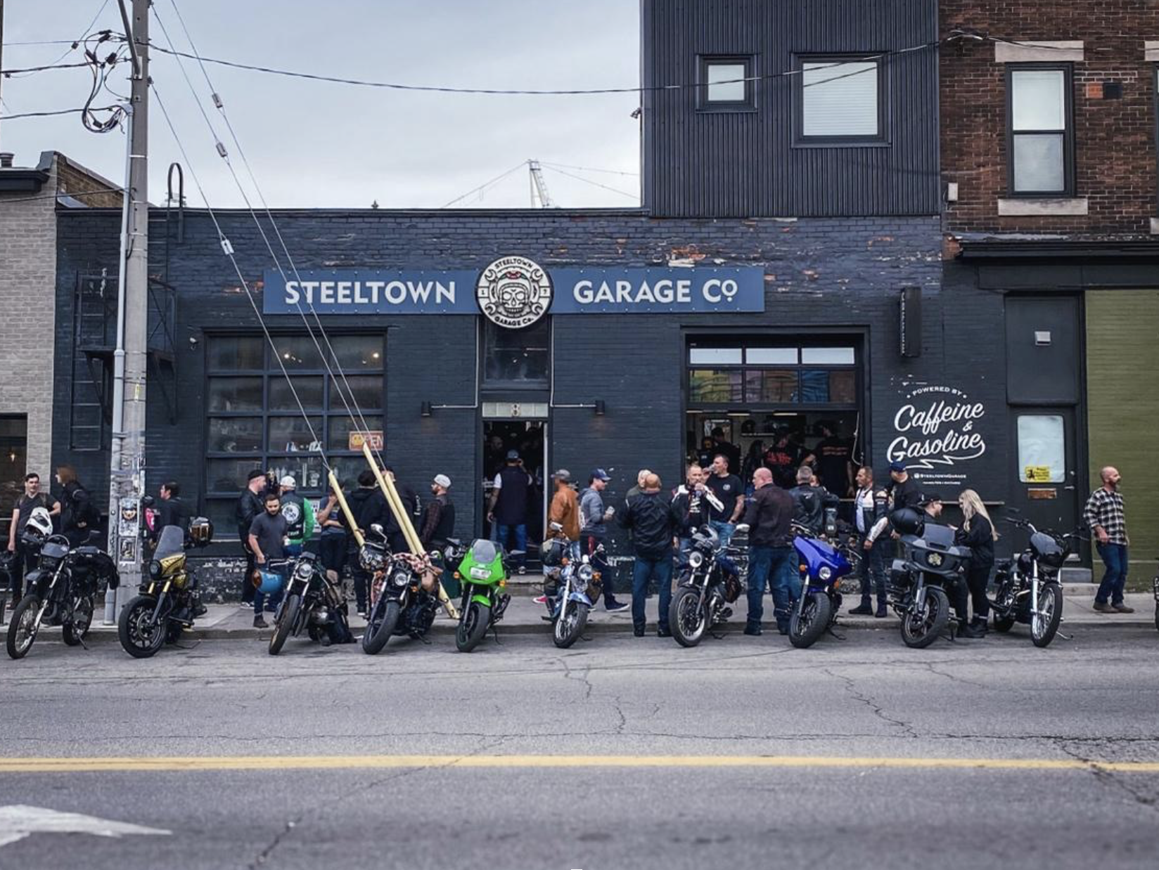 A row of parked motorcycles on the street in front of a dark blue business with the sign "Steeltown Garage". Riders are talking in a group on the sidewalk.