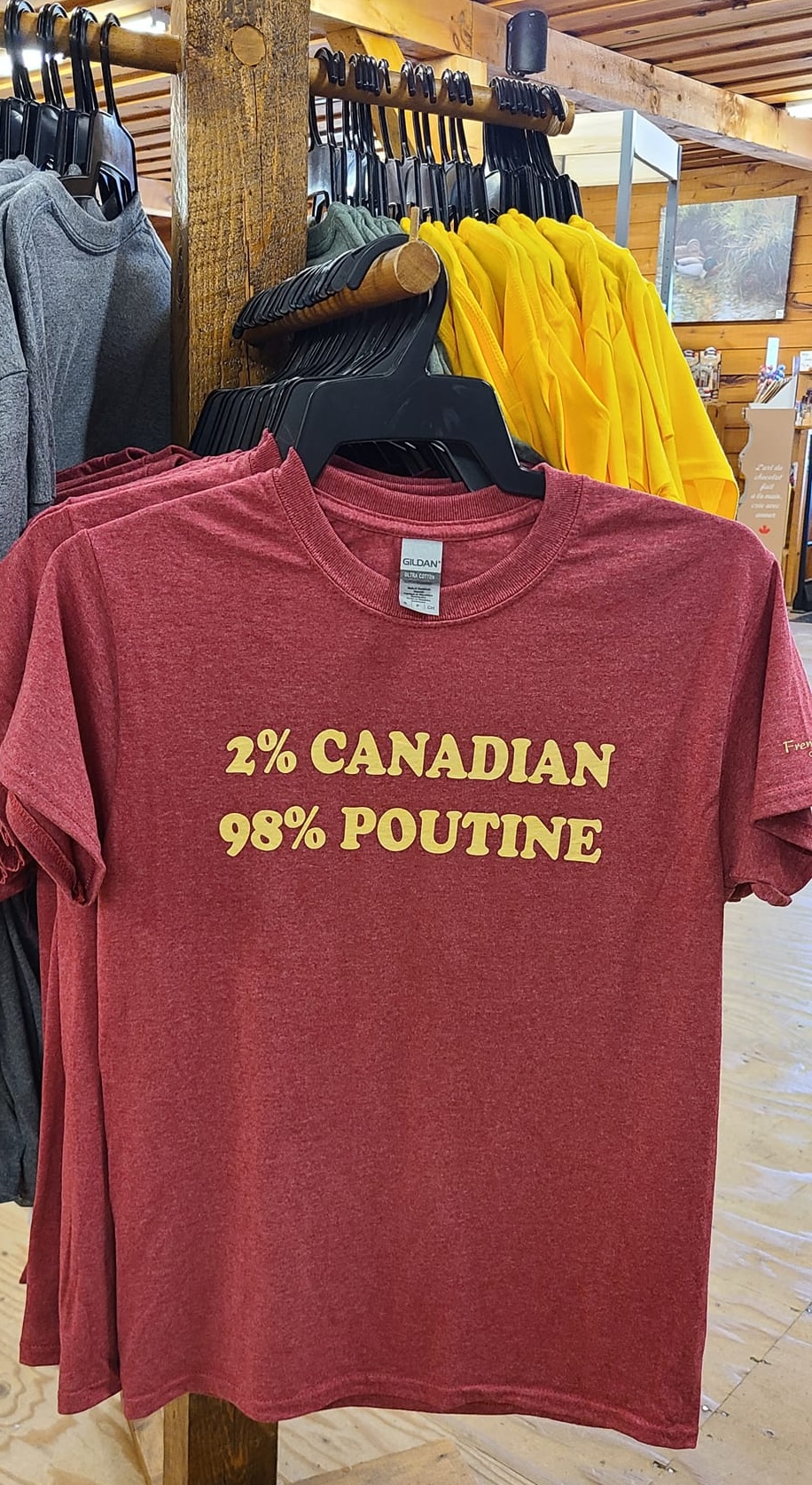 a rack of colourful T-shirts on display in the French River Trading Post. The dark red shirts facing the camera read "2% Canadian, 98% Poutine".  