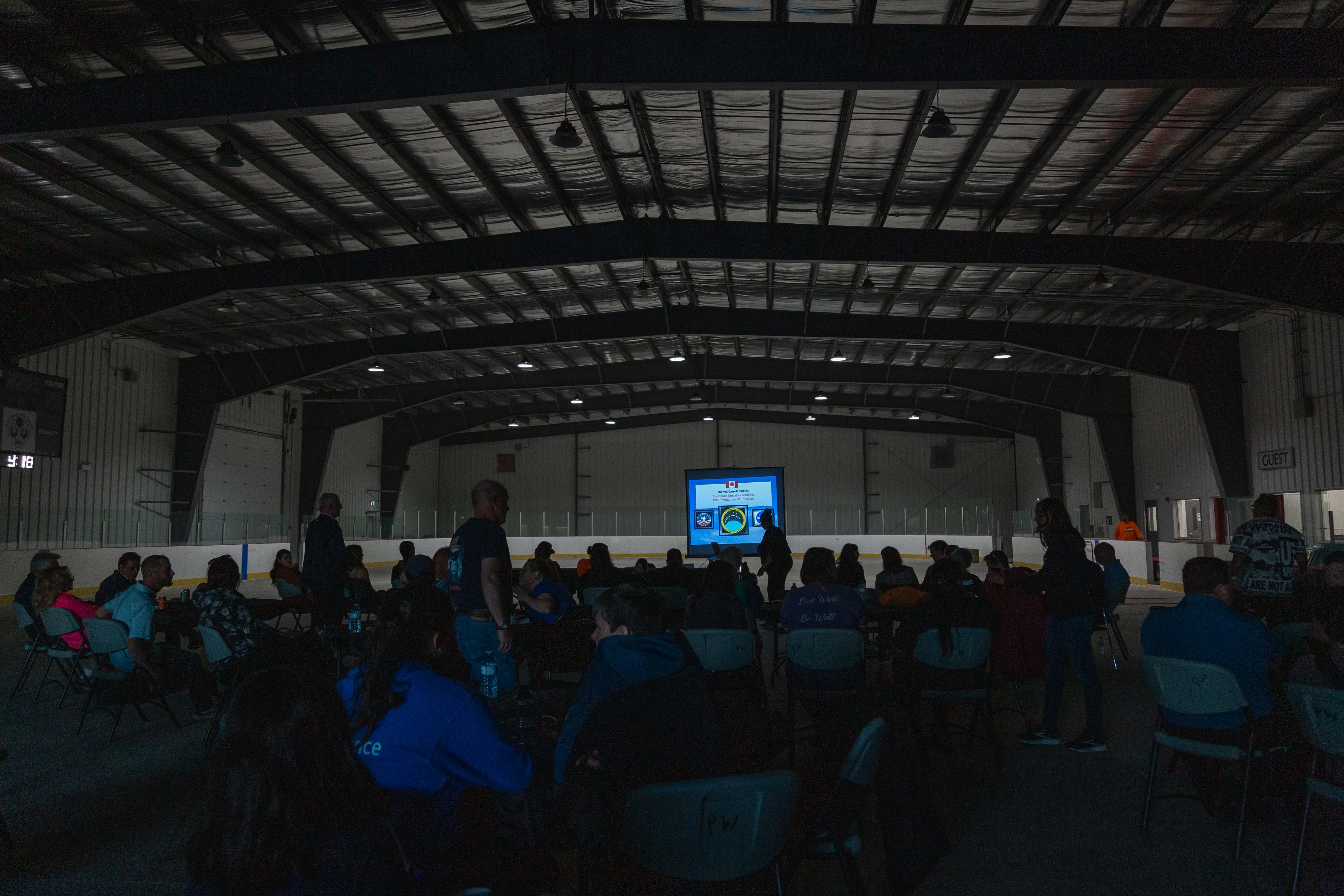 students sitting in a crowd in a darkened arena, watching an educational presentation on an illuminated screen at the Anangokaa Festival.