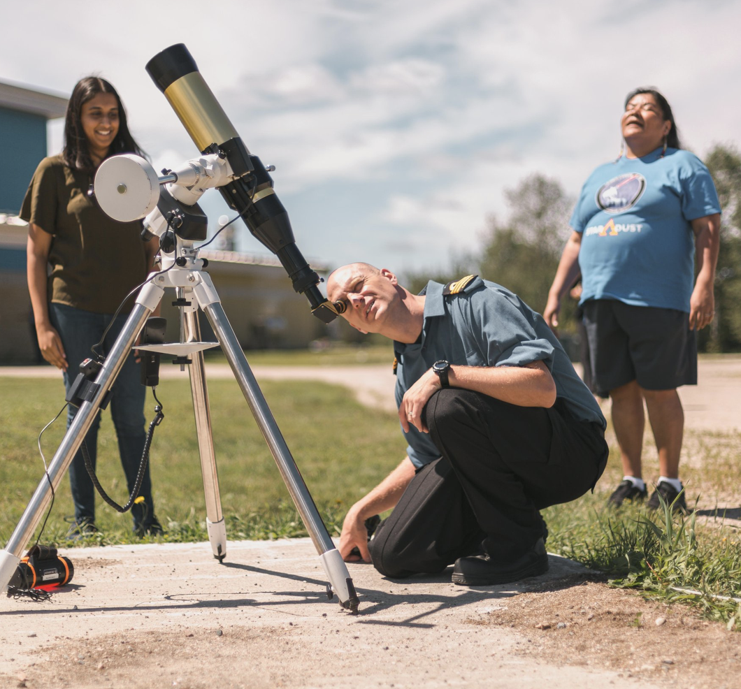 2 smiling students watch as an instructor with the Anangokaa Festival looks up at the sky through a telescope.