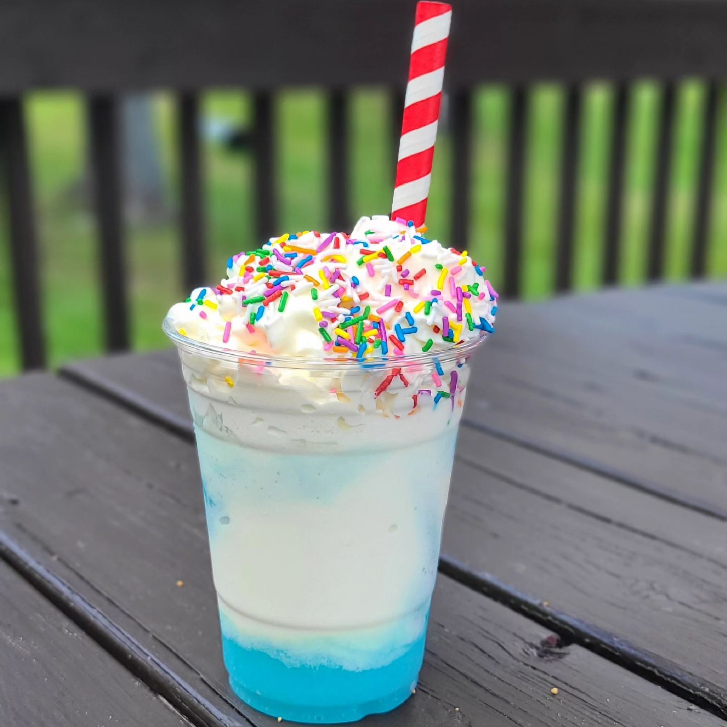 The Blue Hound Freeze from French River Trading Post; a milkshake with bright blue syrup on the bottom topped with whipped cream and rainbow sprinkles. It is sitting on a wooden picnic table with green grass in the background.