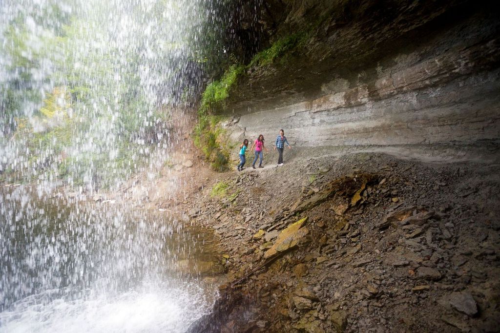 Bridal Veil Falls; a few people walk in a line along the steep, smooth rockface behind the curtain of water of the falls.  