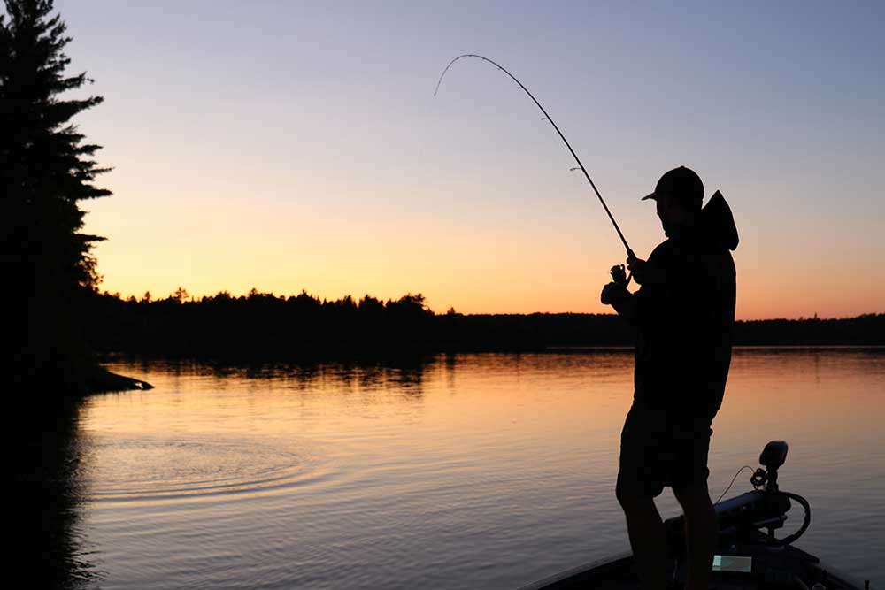 a man standing at a lake in a forest, reeling in a fish. He is silhouetted black against an orange sunset. 