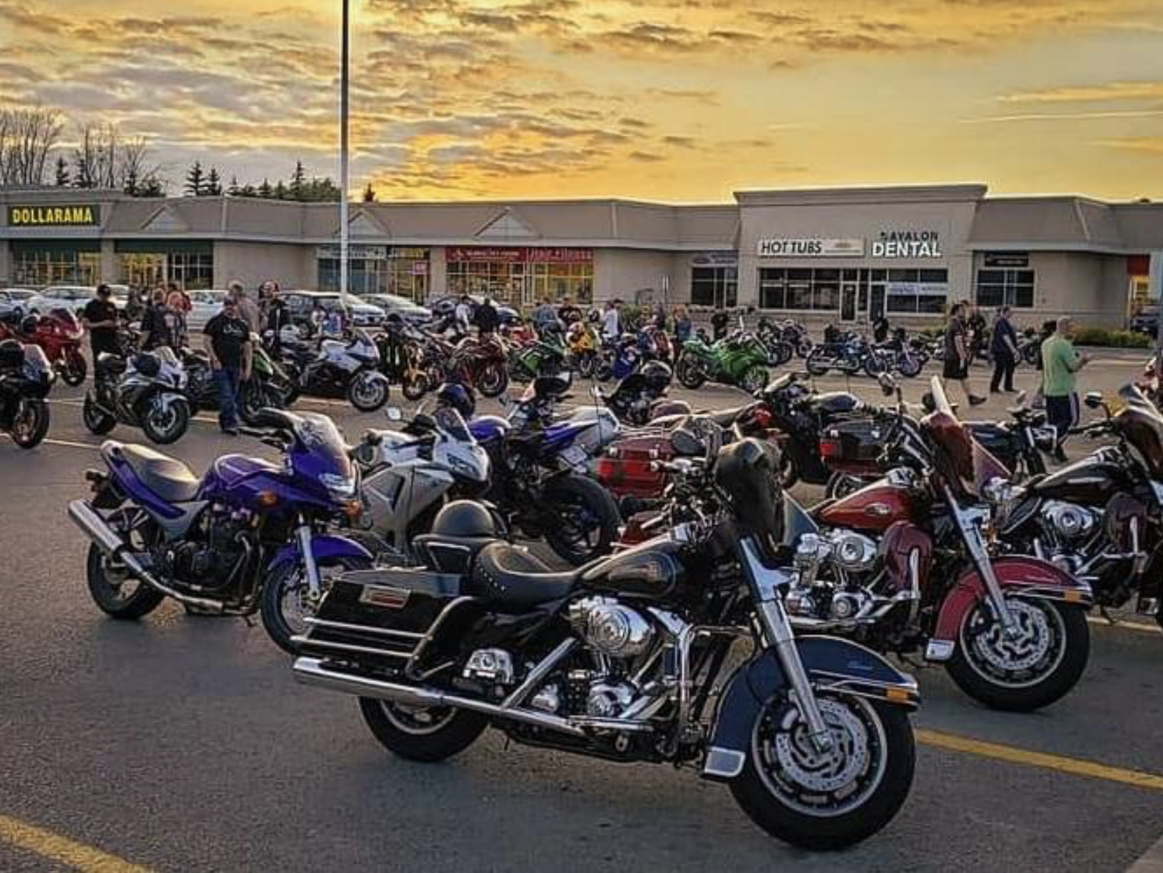 A strip mall parking lot filled with rows of parked motorcycles and groups of people standing around visiting under a golden sunset on a summer evening. 