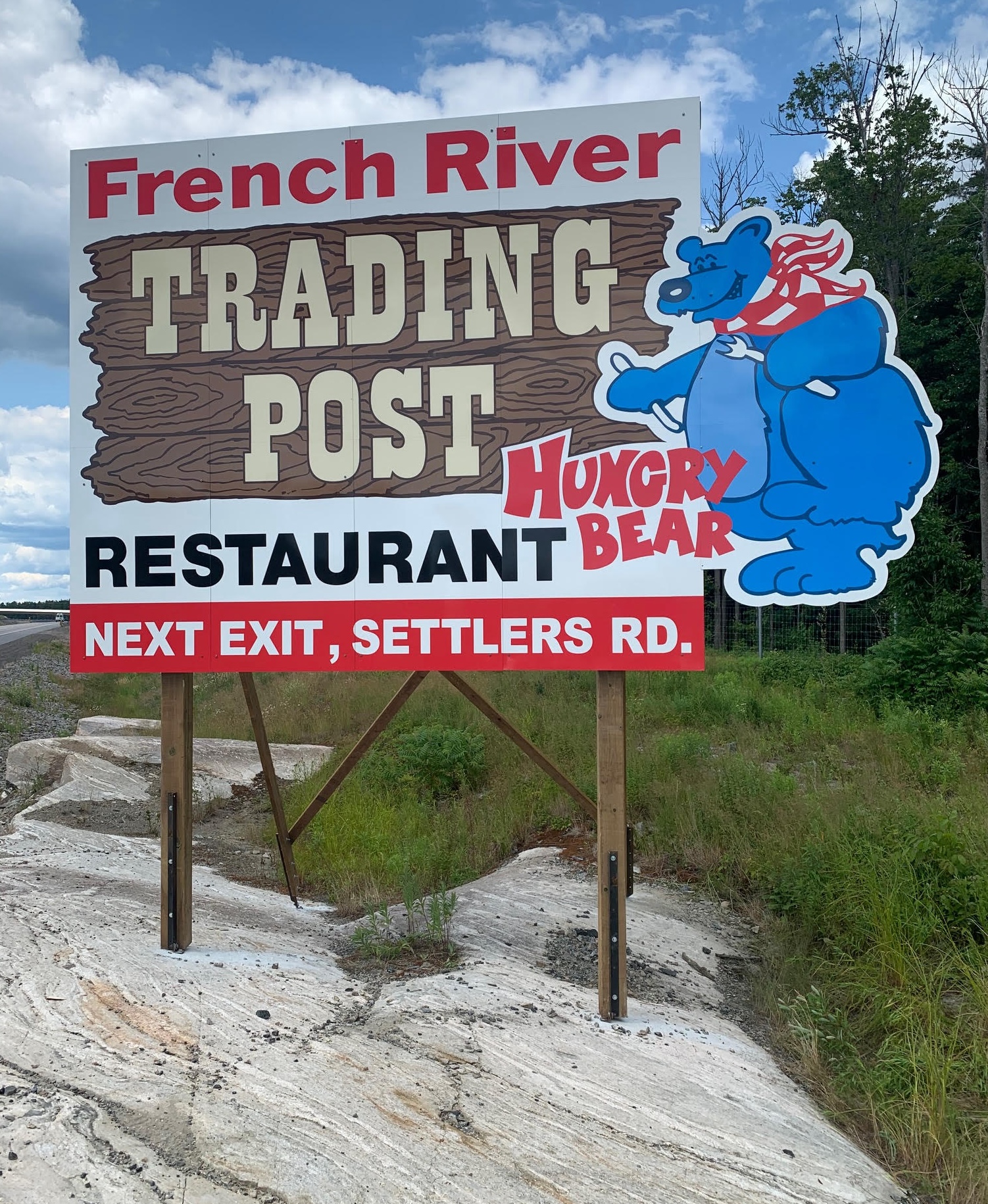 A large sign standing at a rocky and forested roadside, advertising the French River Trading Post and Hungry Bear Restaurant. The sign features a blue bear with a napkin around its neck, holding a knife and fork. 