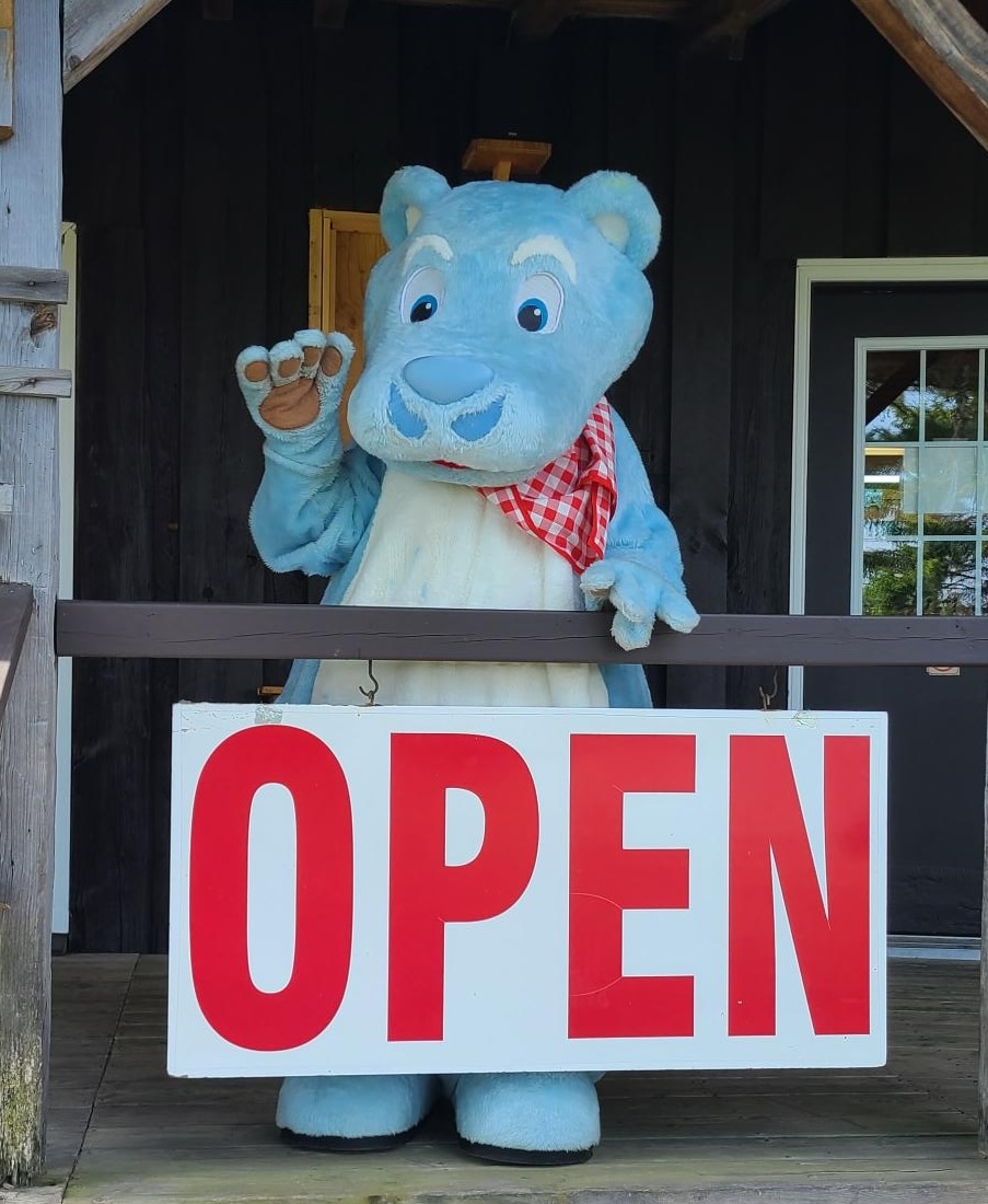 a blue bear mascot with a moustache and a red bandana waves from behind a large "OPEN" sign on a wooden railing in front of the French River Trading Post.